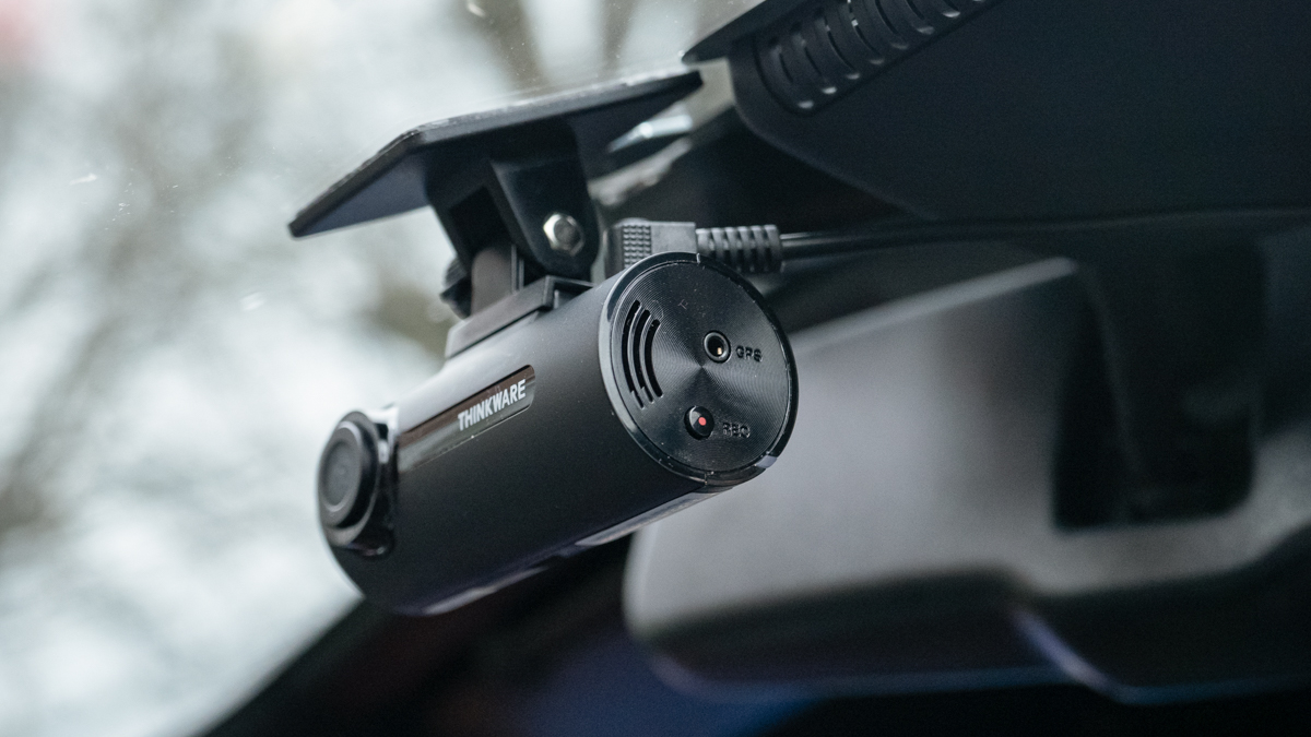 Compact black dash cam mounted on a windshield near the rearview mirror with bare trees outside