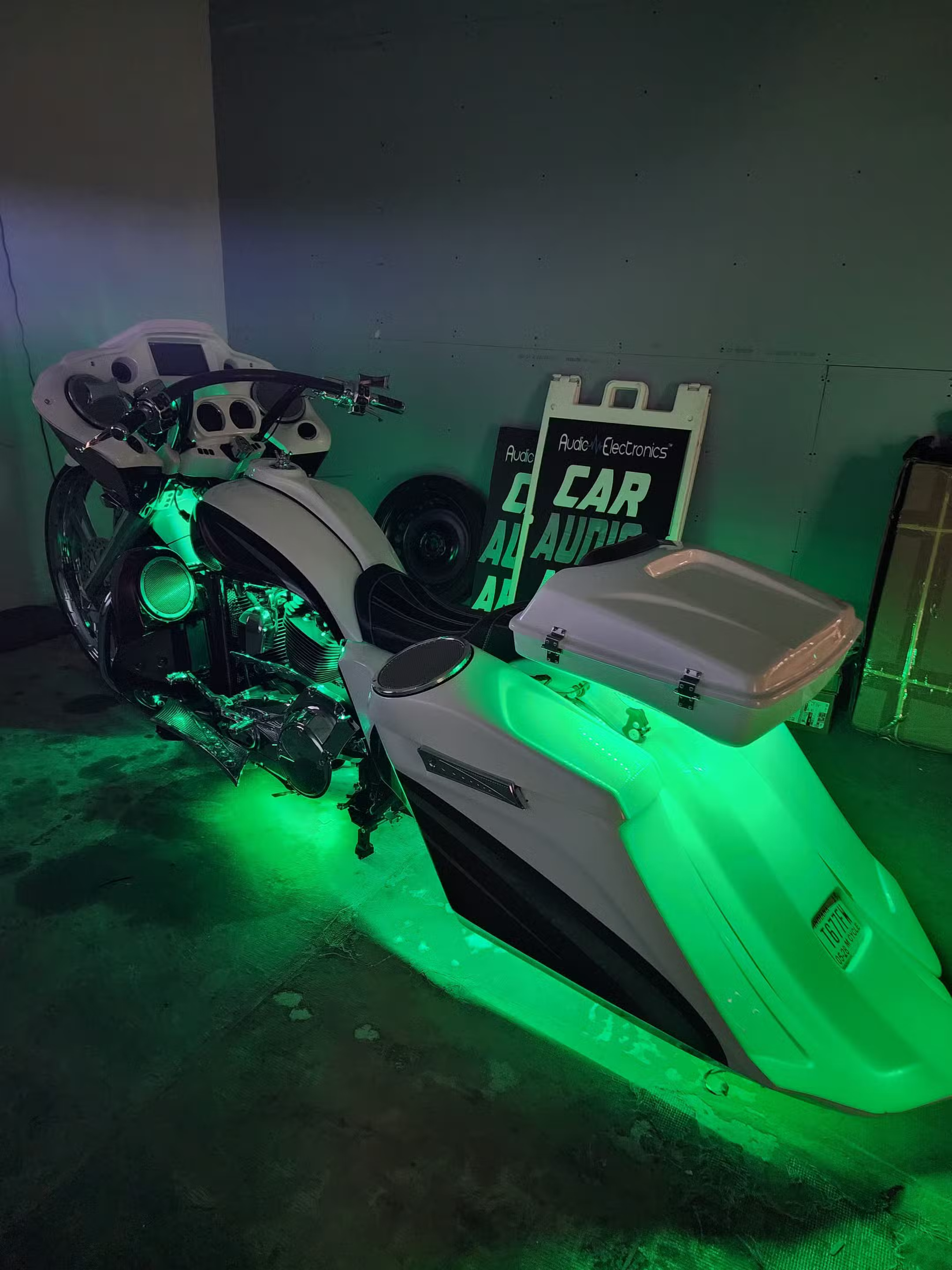 White custom bagger motorcycle glowing with green LED underglow beside a car audio shop sign.