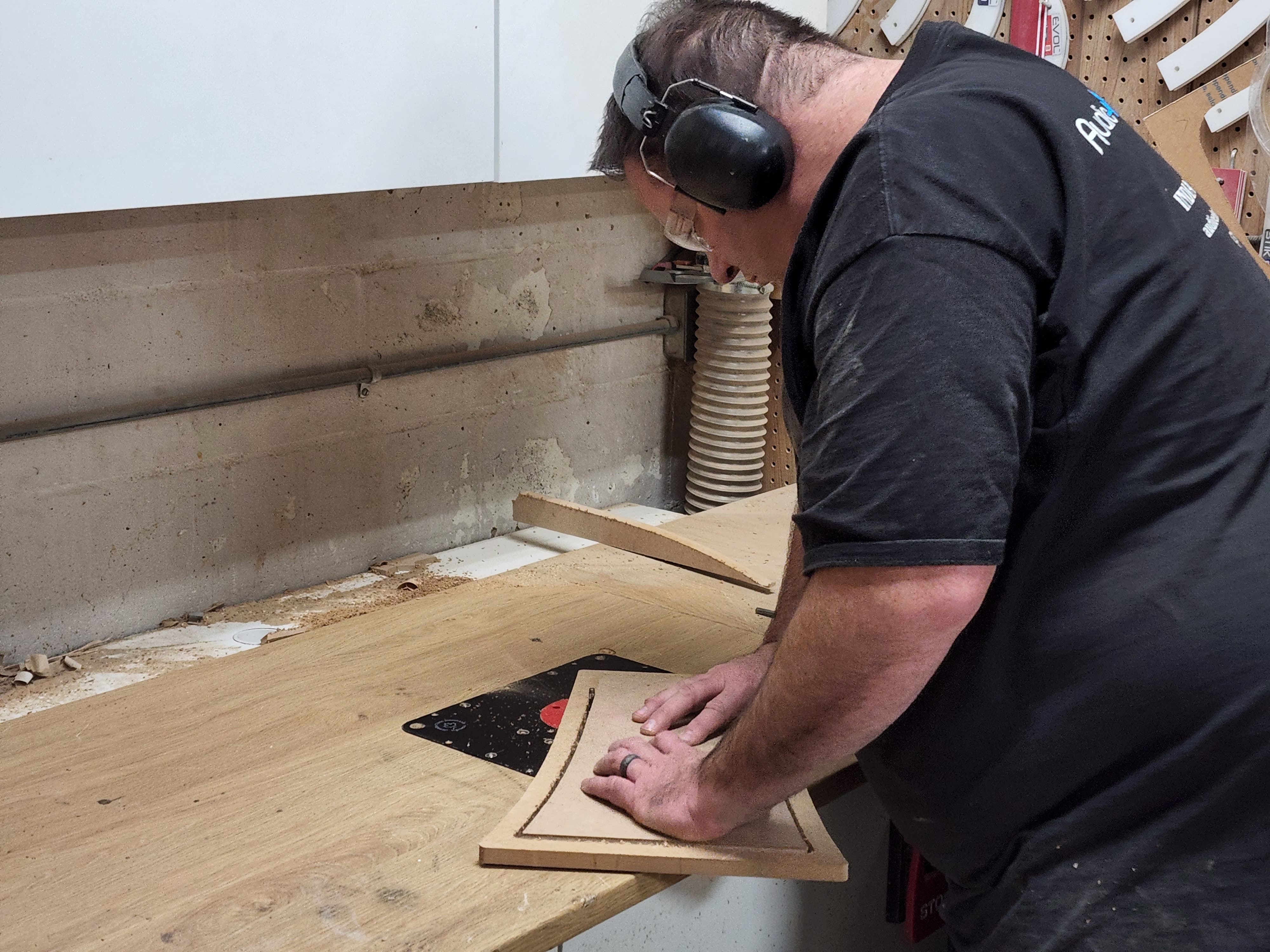 Installer wearing ear protection shapes a curved wooden speaker baffle on a dusty fabrication bench.