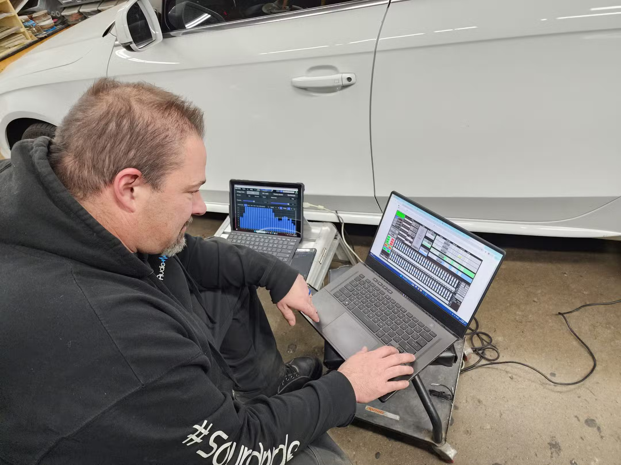Installer kneeling next to a white car adjusting tuning software on a laptop while a tablet shows a frequency graph