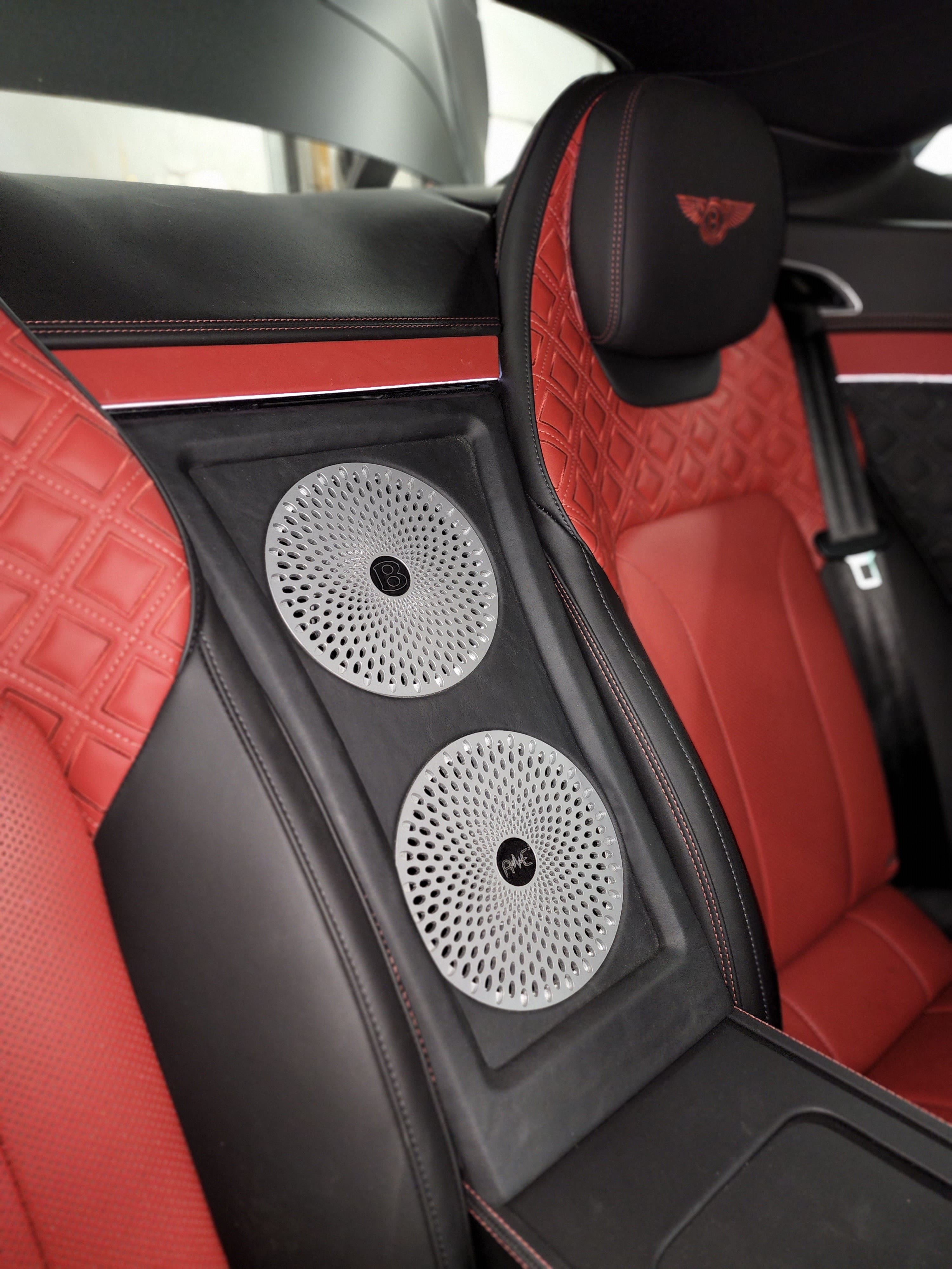 Two large mesh grille speakers mounted in a black panel between red quilted leather rear seats of a luxury coupe.