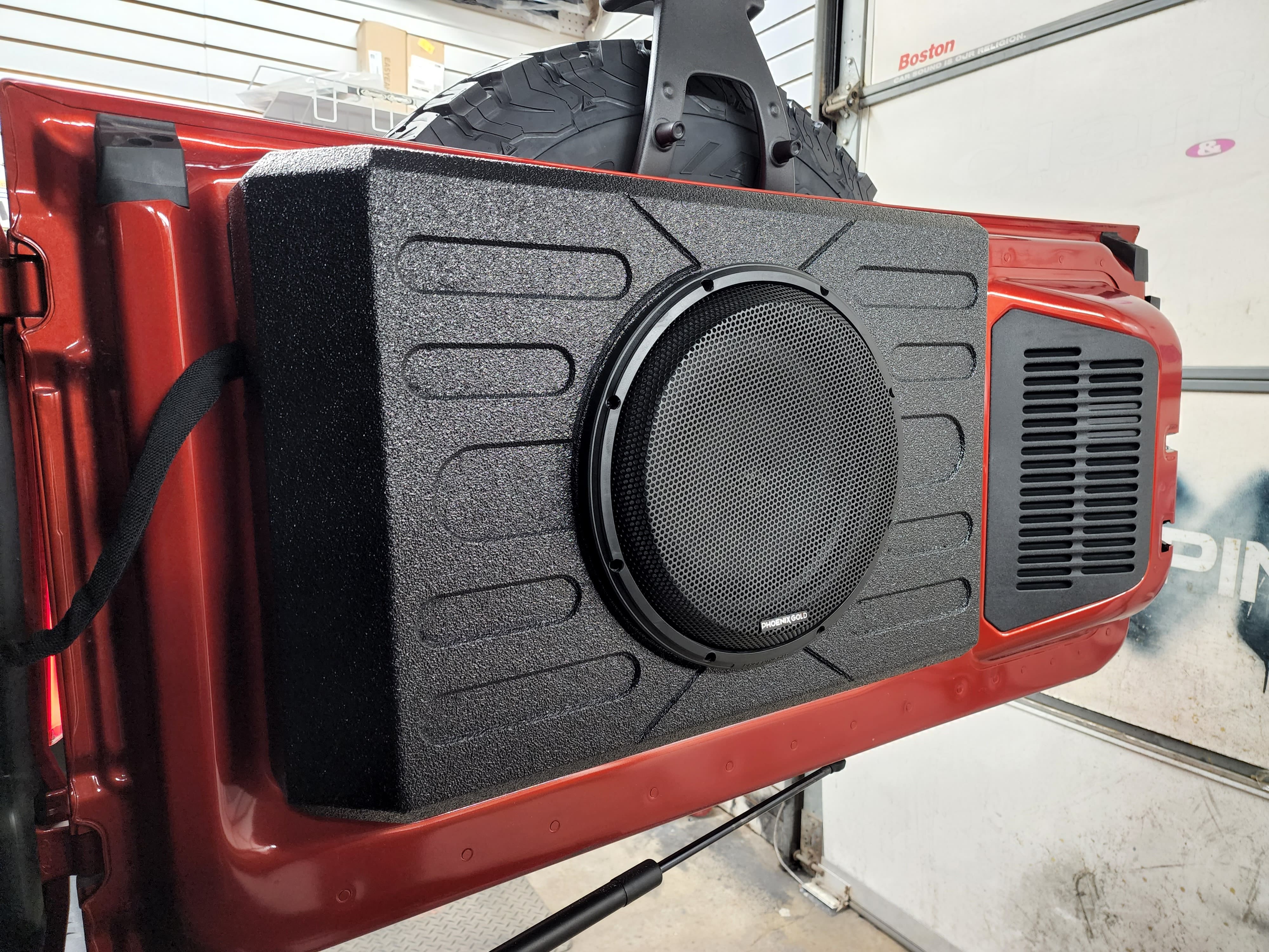 Carpeted single subwoofer enclosure with a mesh grille resting on the open tailgate of a red Jeep inside a shop bay.