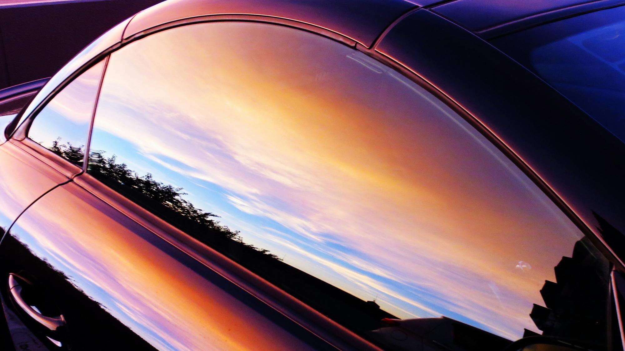 Orange sunset sky and a tree line reflected across the tinted side window of a coupe.