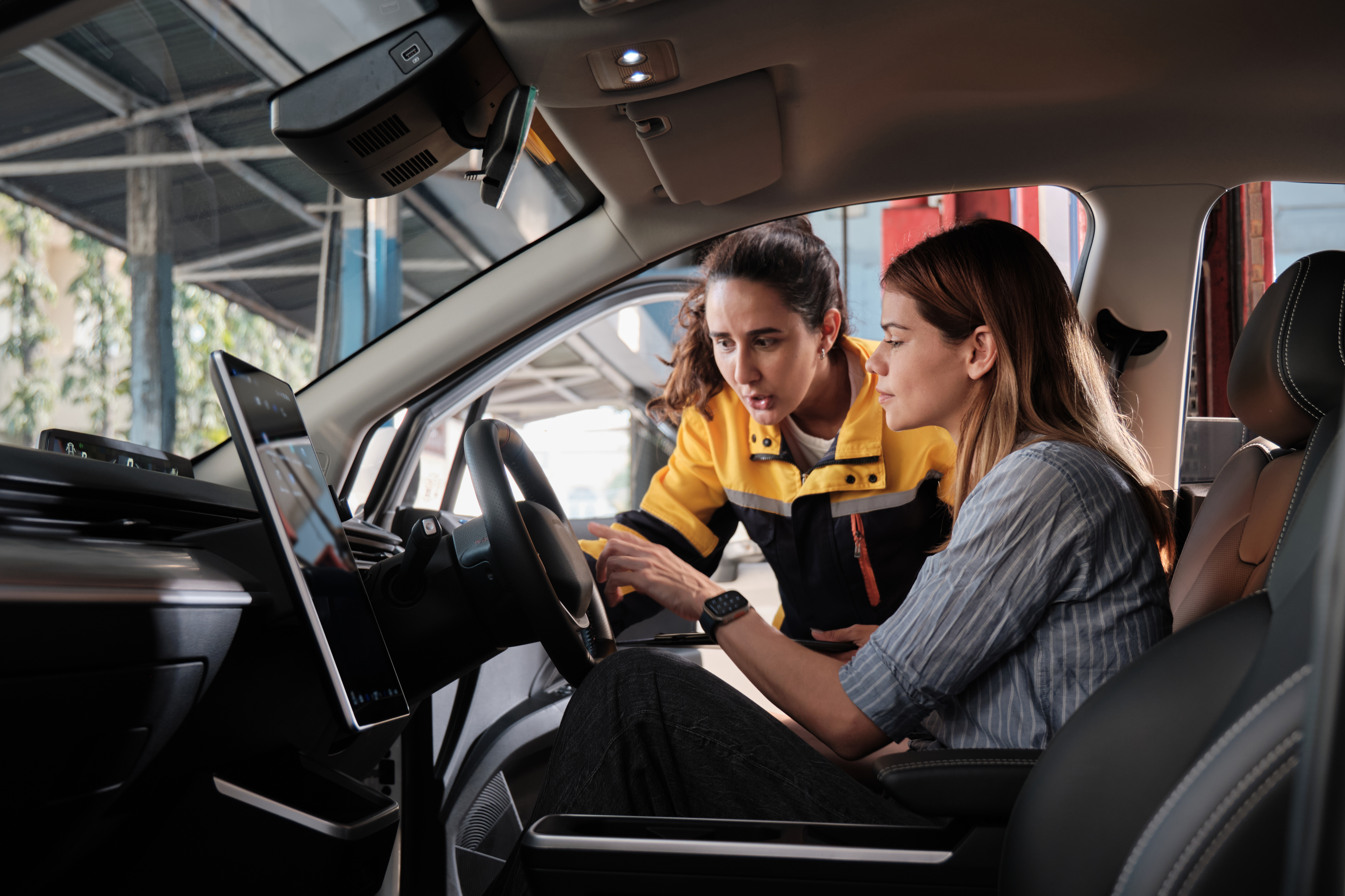 Service adviser in a work jacket leans into a car to show a customer the dashboard touchscreen