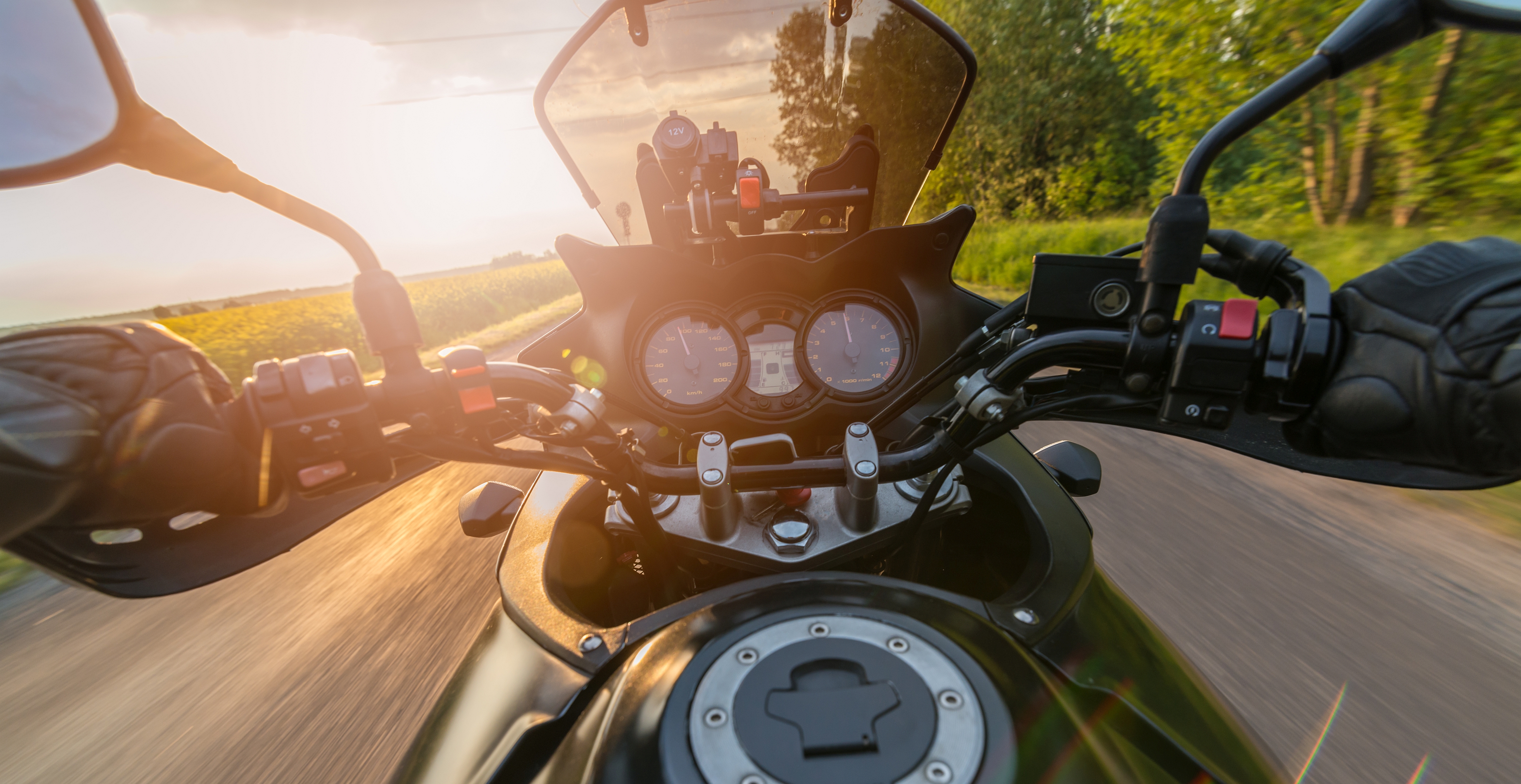 Rider's point of view over a motorcycle's handlebars and gauges on a sunlit country road