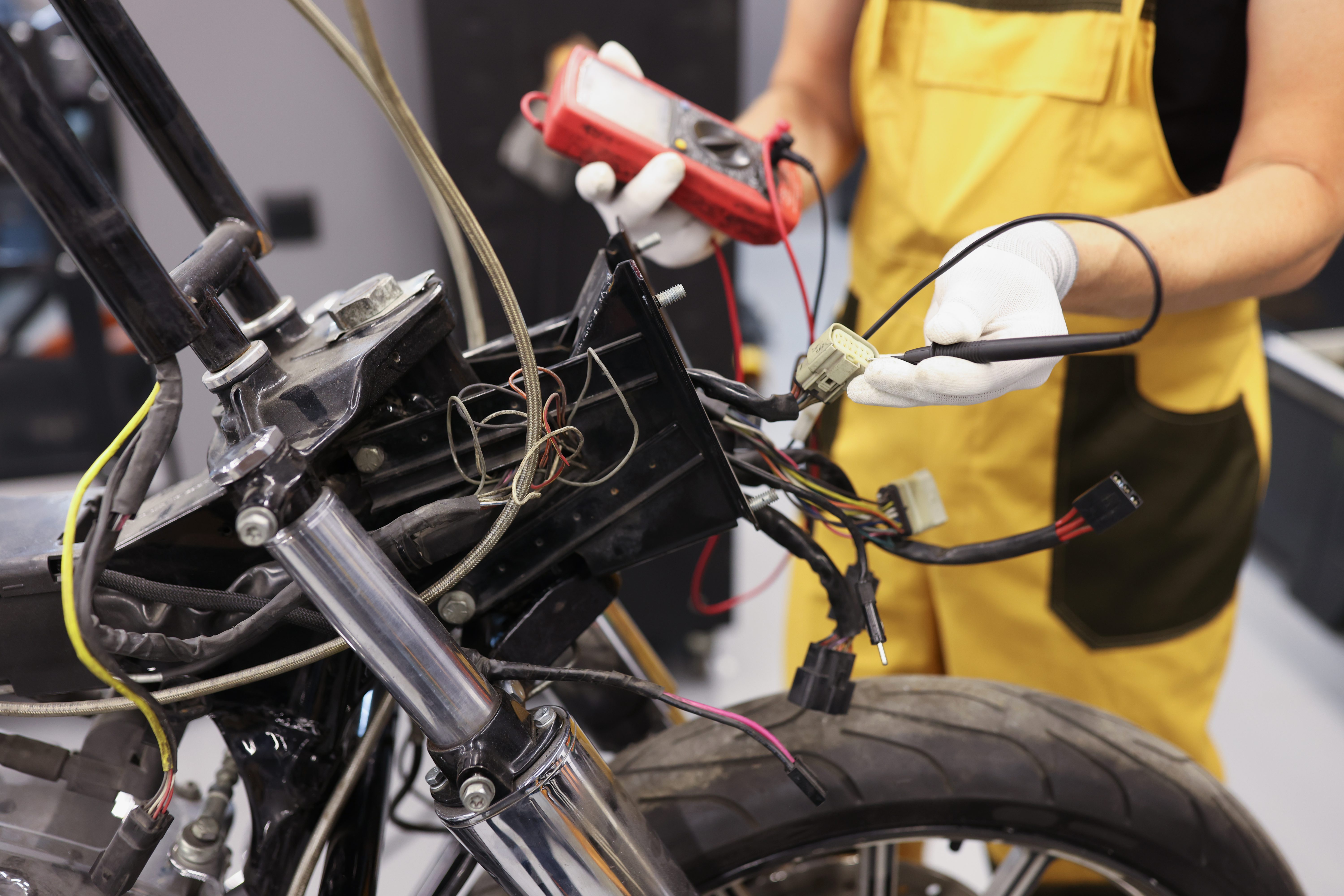 Technician in yellow overalls using a multimeter to check voltage on a motorcycle's front wiring harness