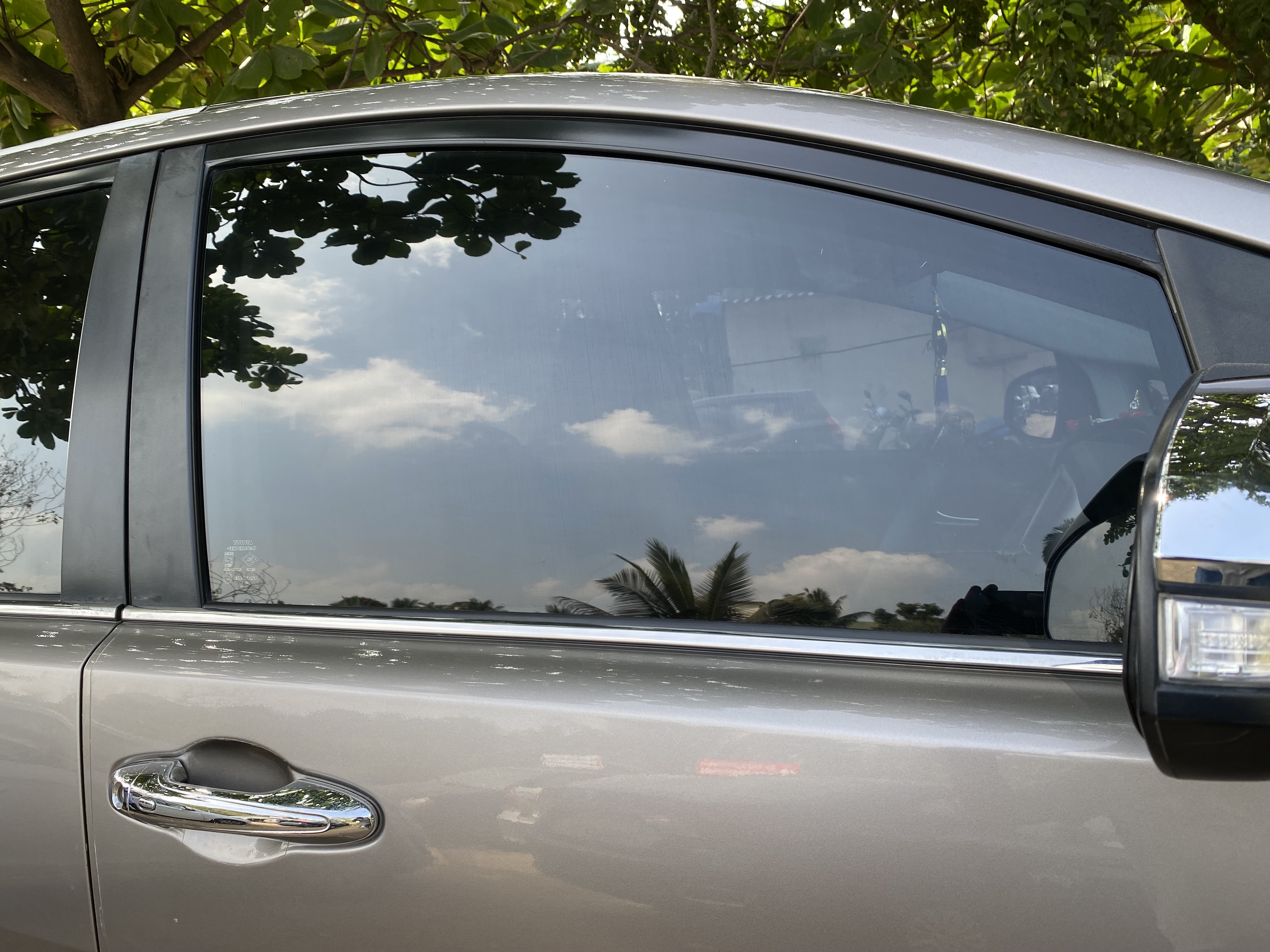 Close view of a silver vehicle's rear door glass with dark tint reflecting sky and trees.