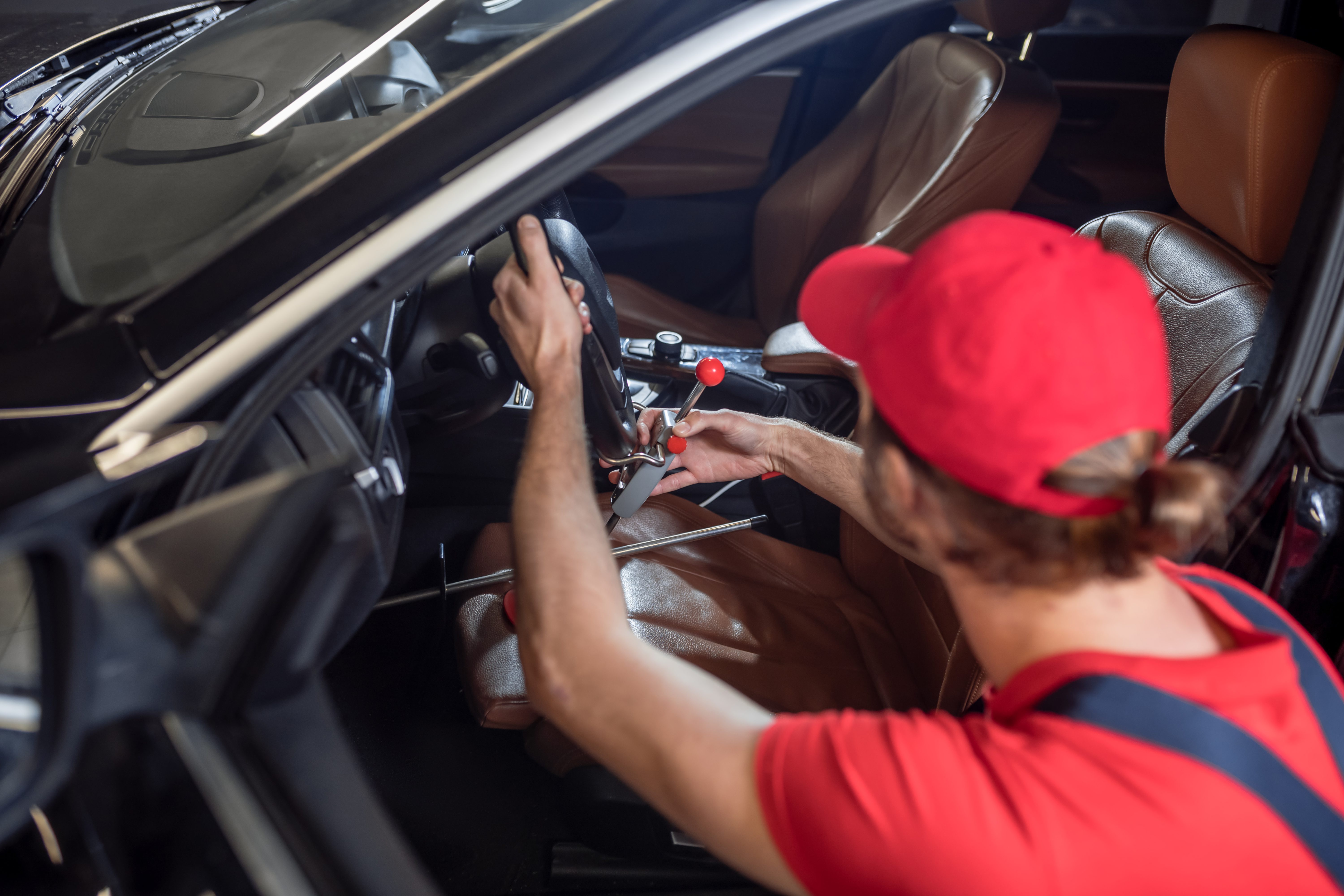 Technician in a red cap and shirt working at the steering column inside a car.
