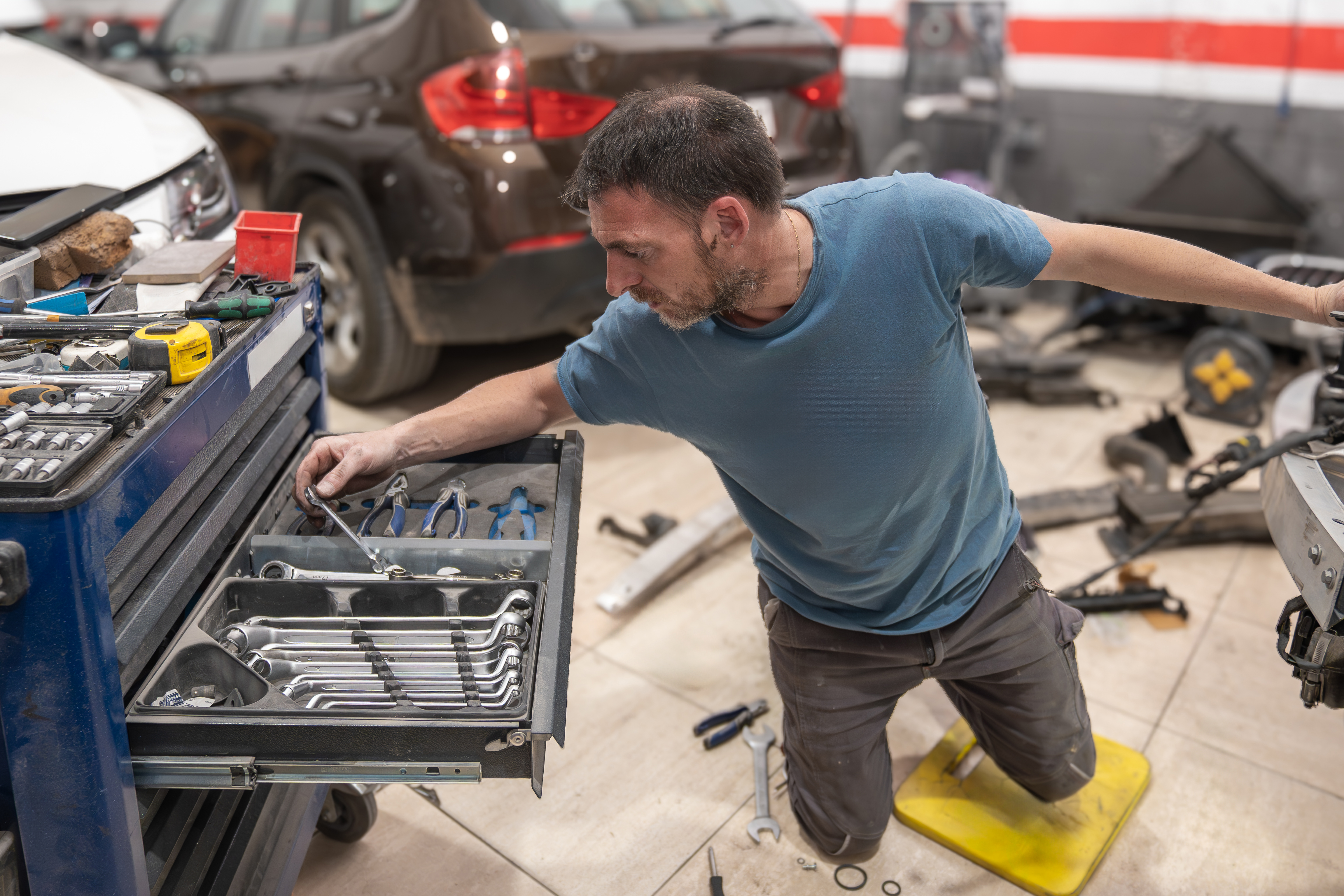 Mechanic kneeling beside a rolling tool chest, choosing wrenches in a busy repair shop.