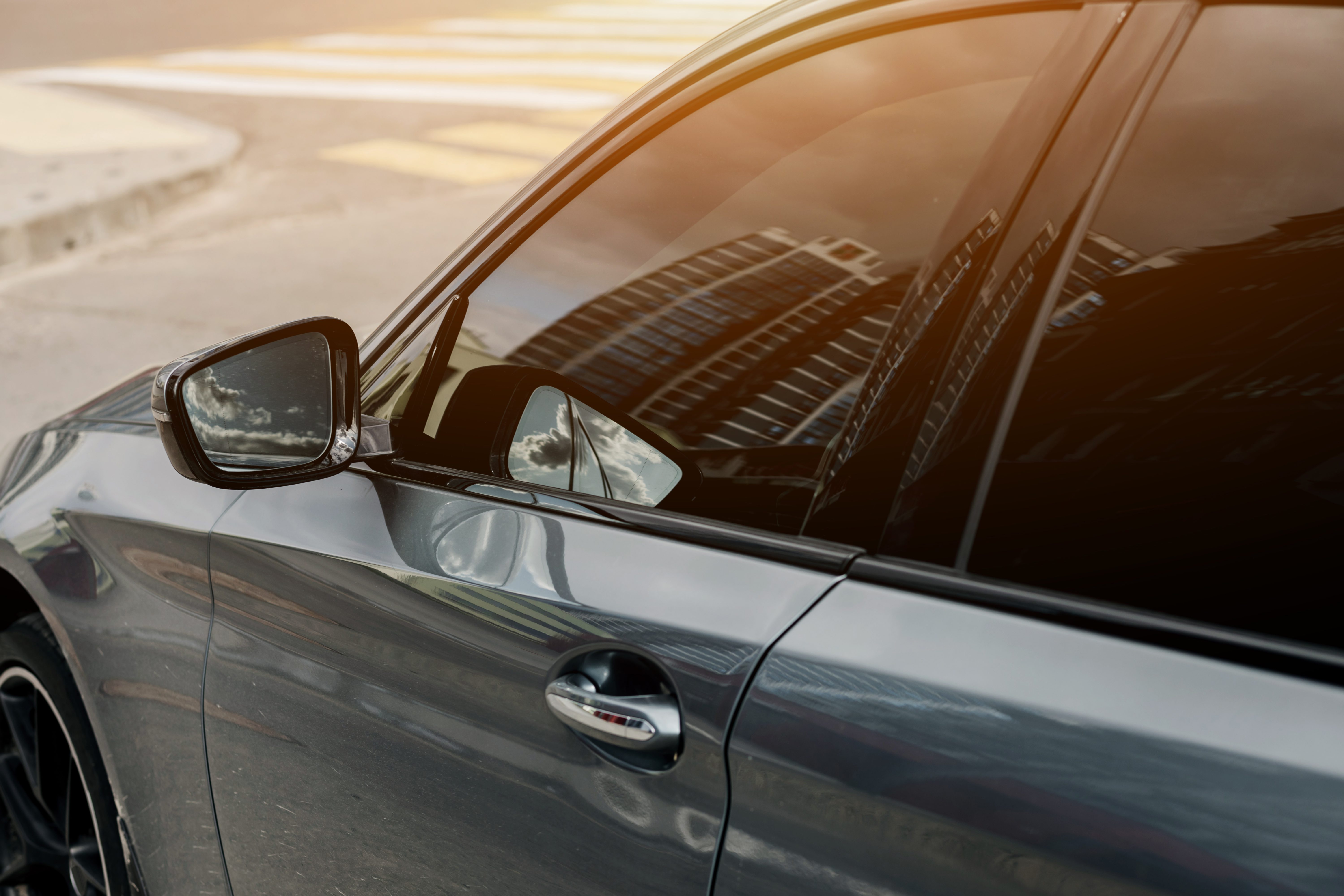 Dark sedan with tinted windows and a side mirror reflecting a tall building in warm sunlight.