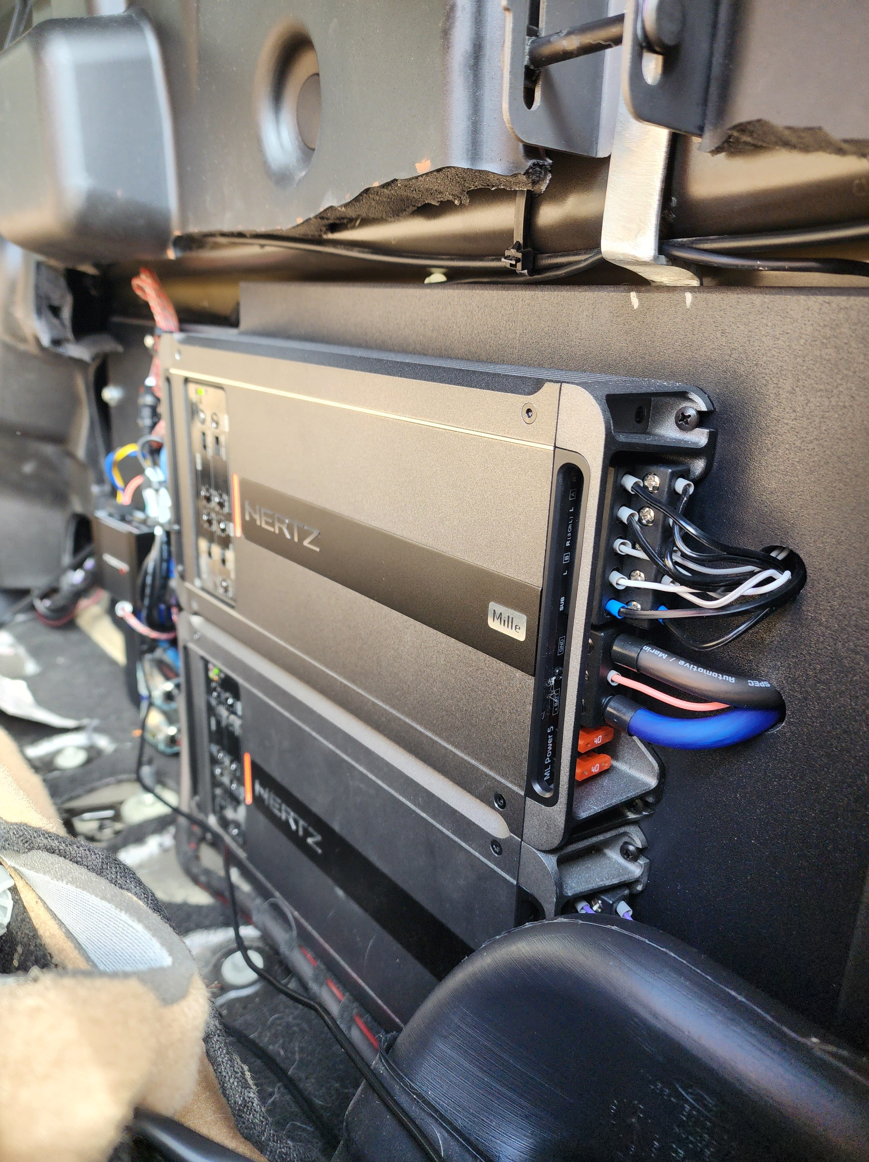 Two black car amplifiers mounted on a panel behind a seat with neatly routed power and speaker wiring.