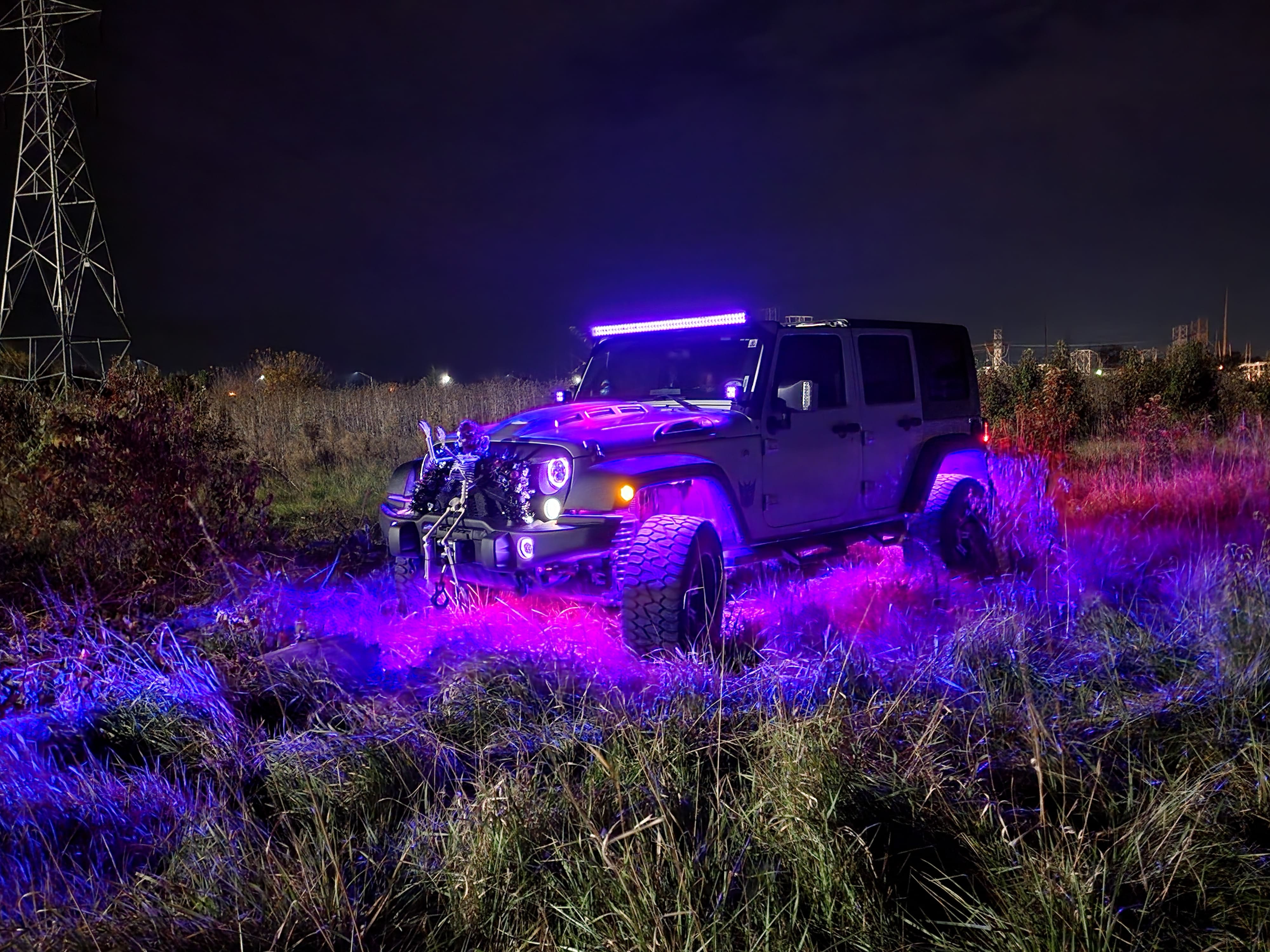 Jeep parked in tall grass at night with a glowing roof light bar and purple underbody lighting.