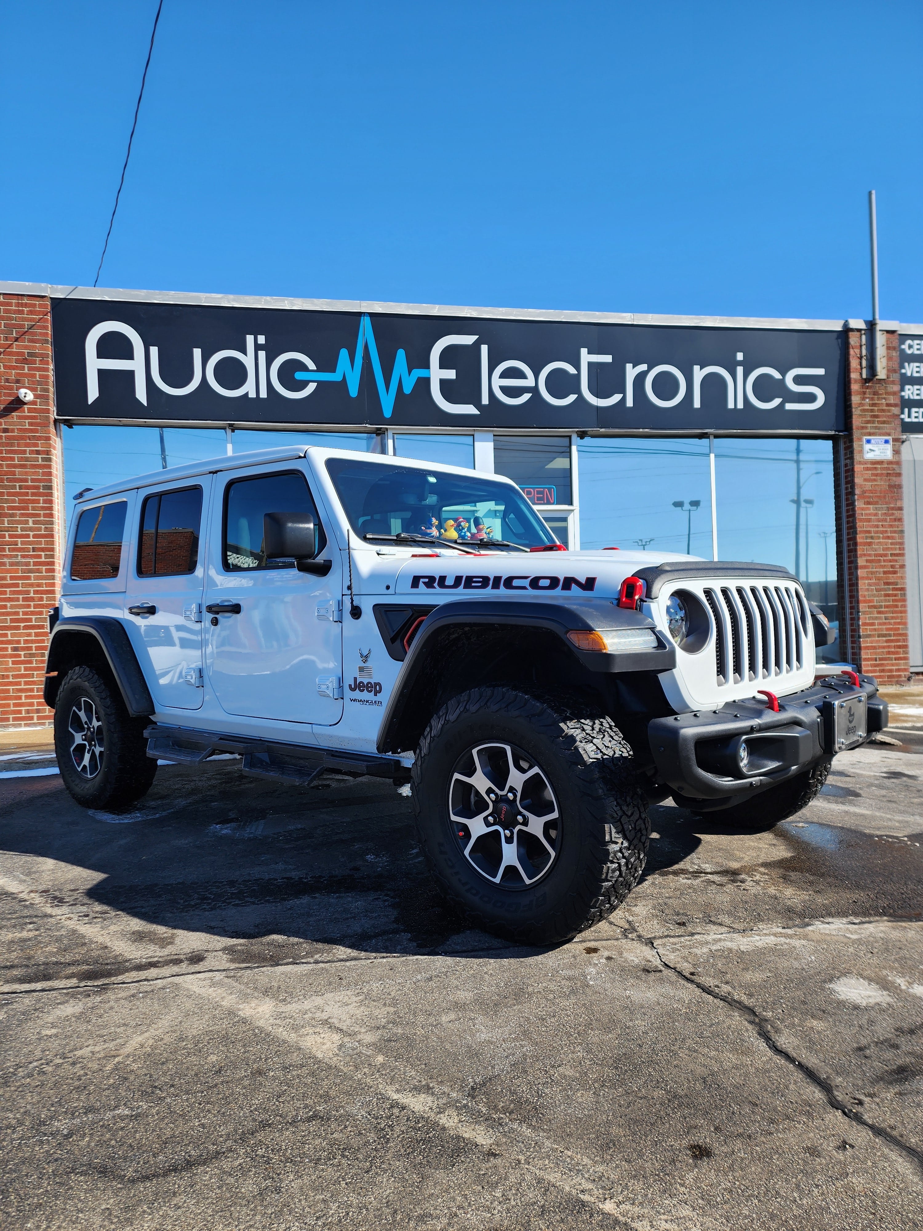 White Jeep Wrangler Rubicon parked outside a brick building with a large Audio Electronics storefront sign.