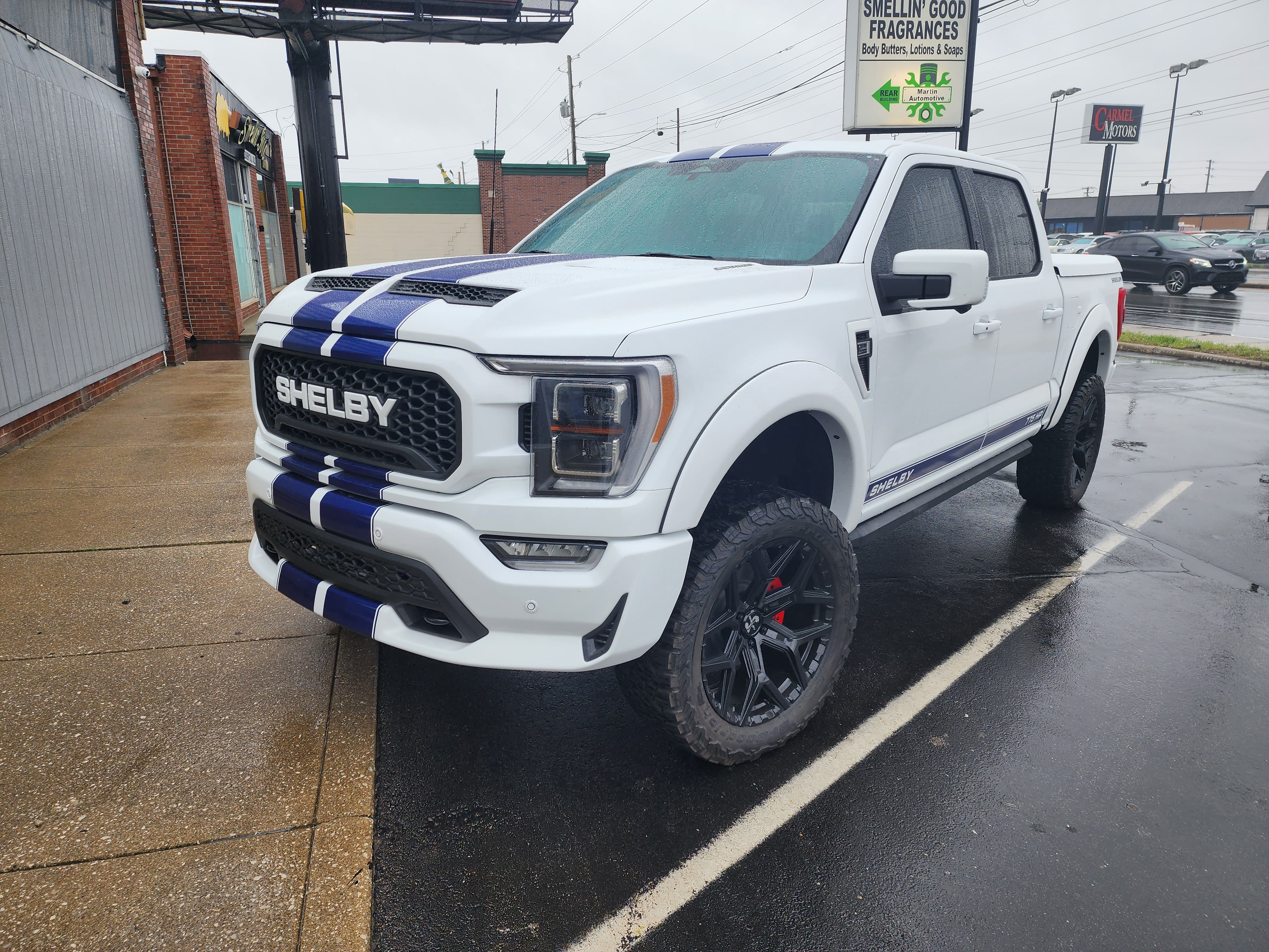 White pickup truck with blue racing stripes, black wheels and knobby all terrain tires parked on wet pavement.