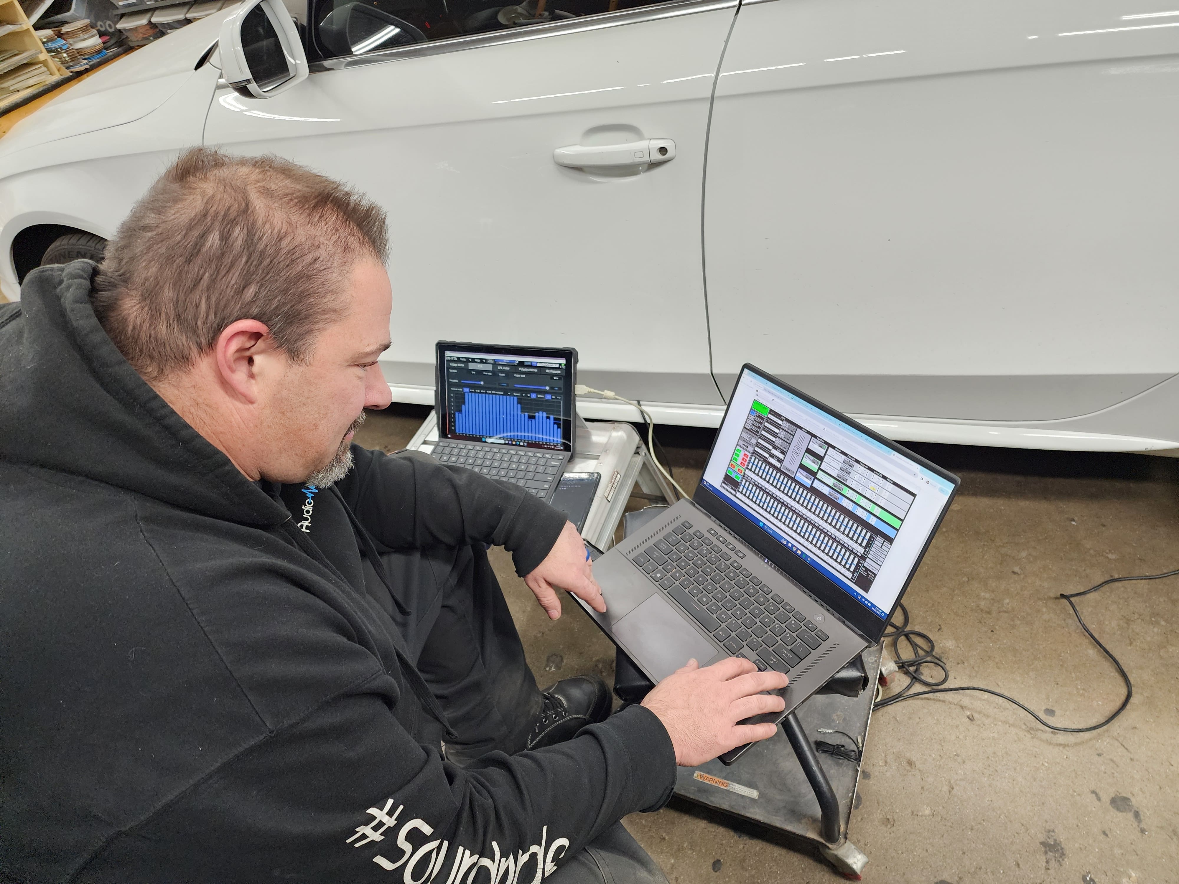 Technician using measurement software on a laptop and tablet to tune a car audio system in a garage.