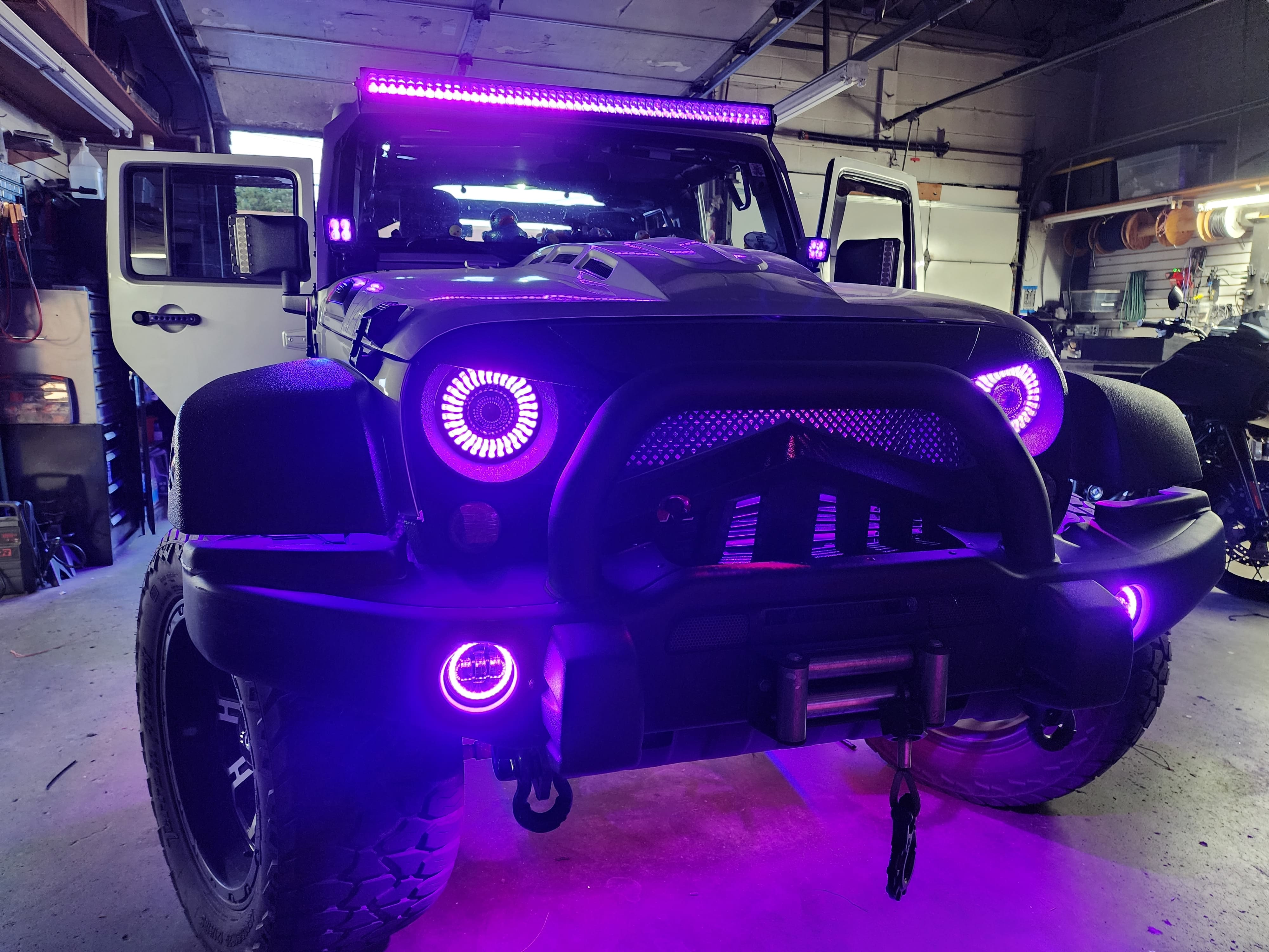 Front of a black Jeep in a garage with purple halo headlights, glowing fog rings and a roof light bar.