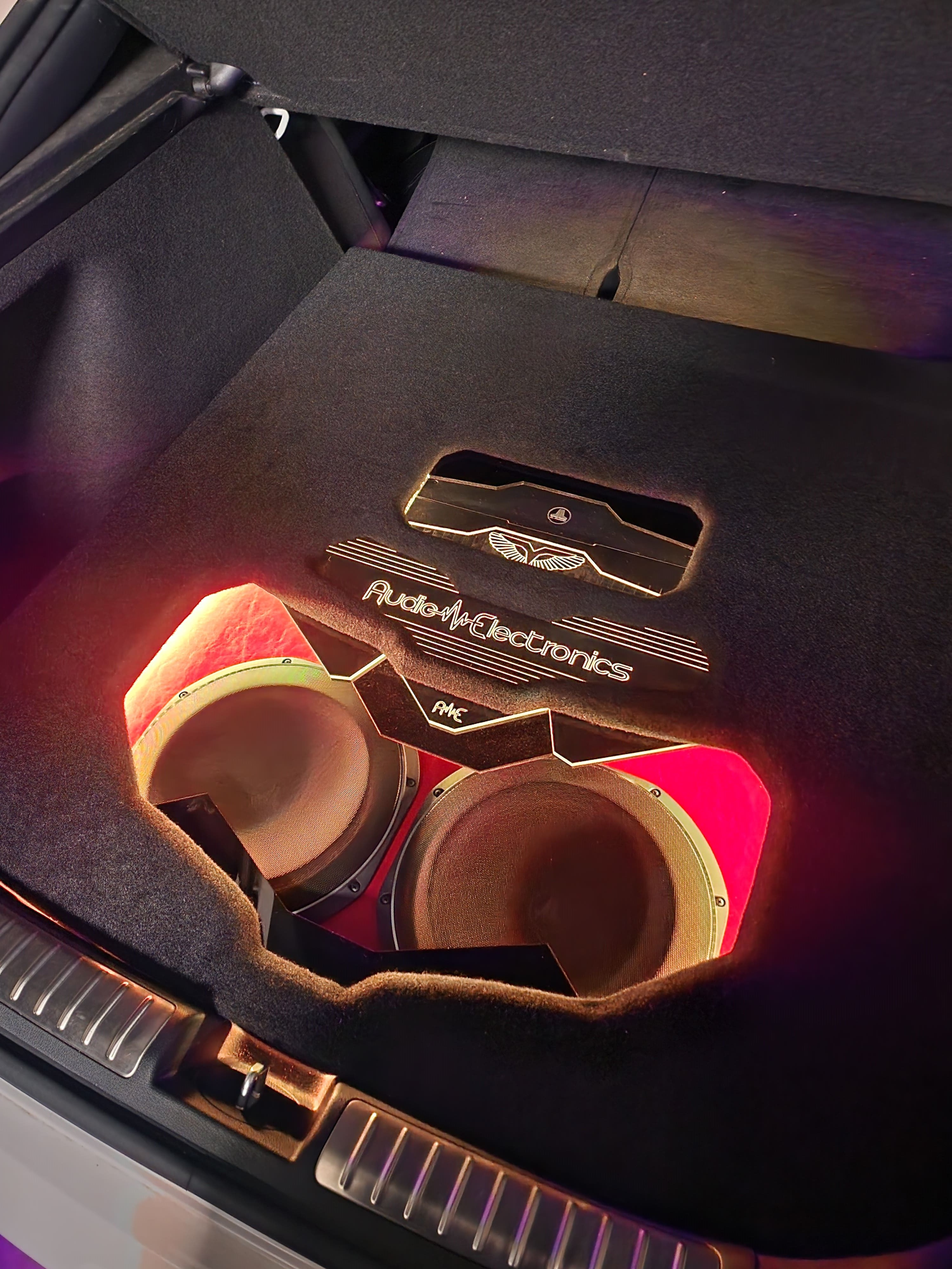 Carpeted trunk enclosure holding two subwoofers behind a lit panel with red accent lighting and a shop logo.
