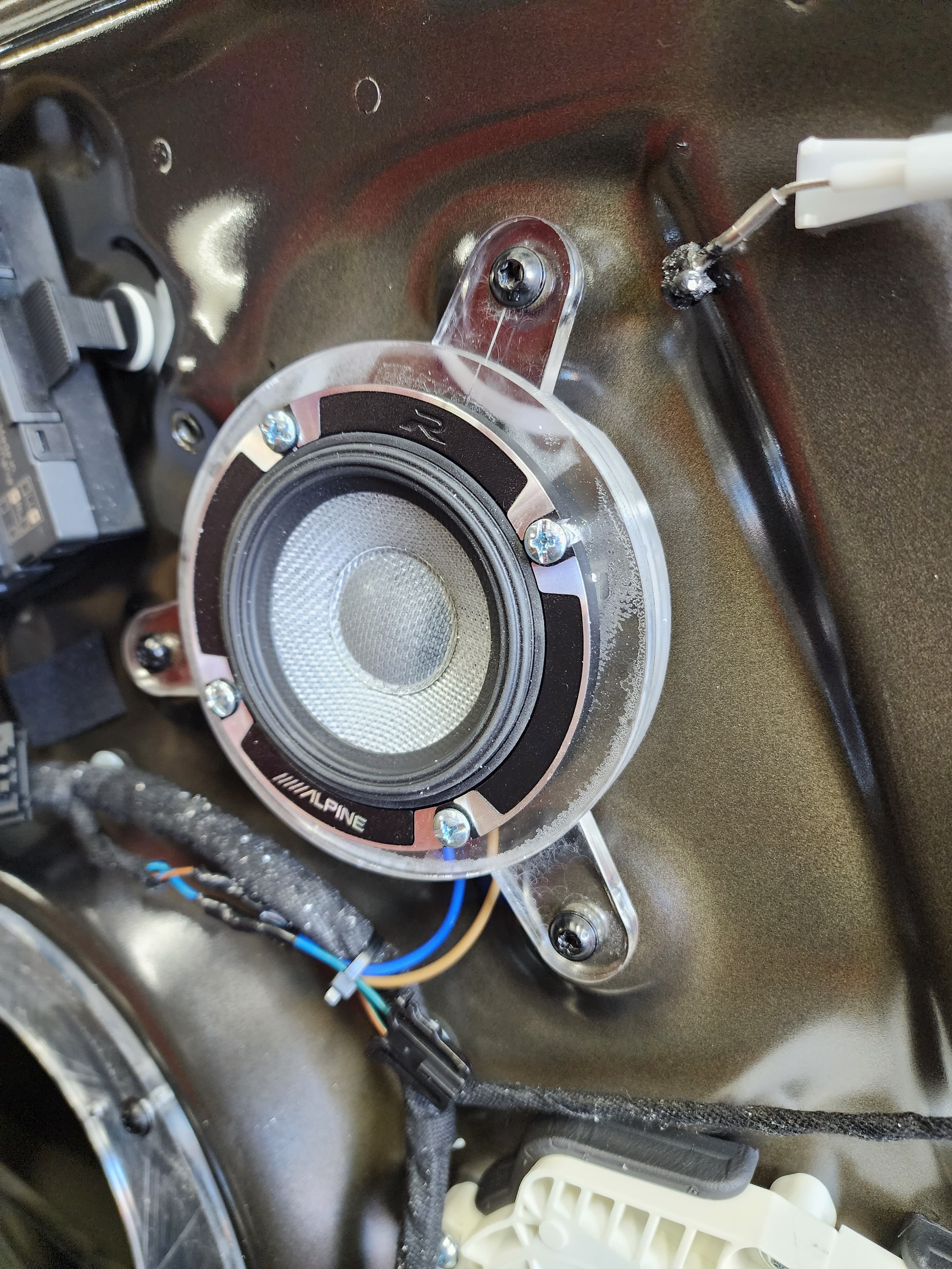 Small component speaker fastened to a clear adapter bracket inside a stripped vehicle door with wiring running past.