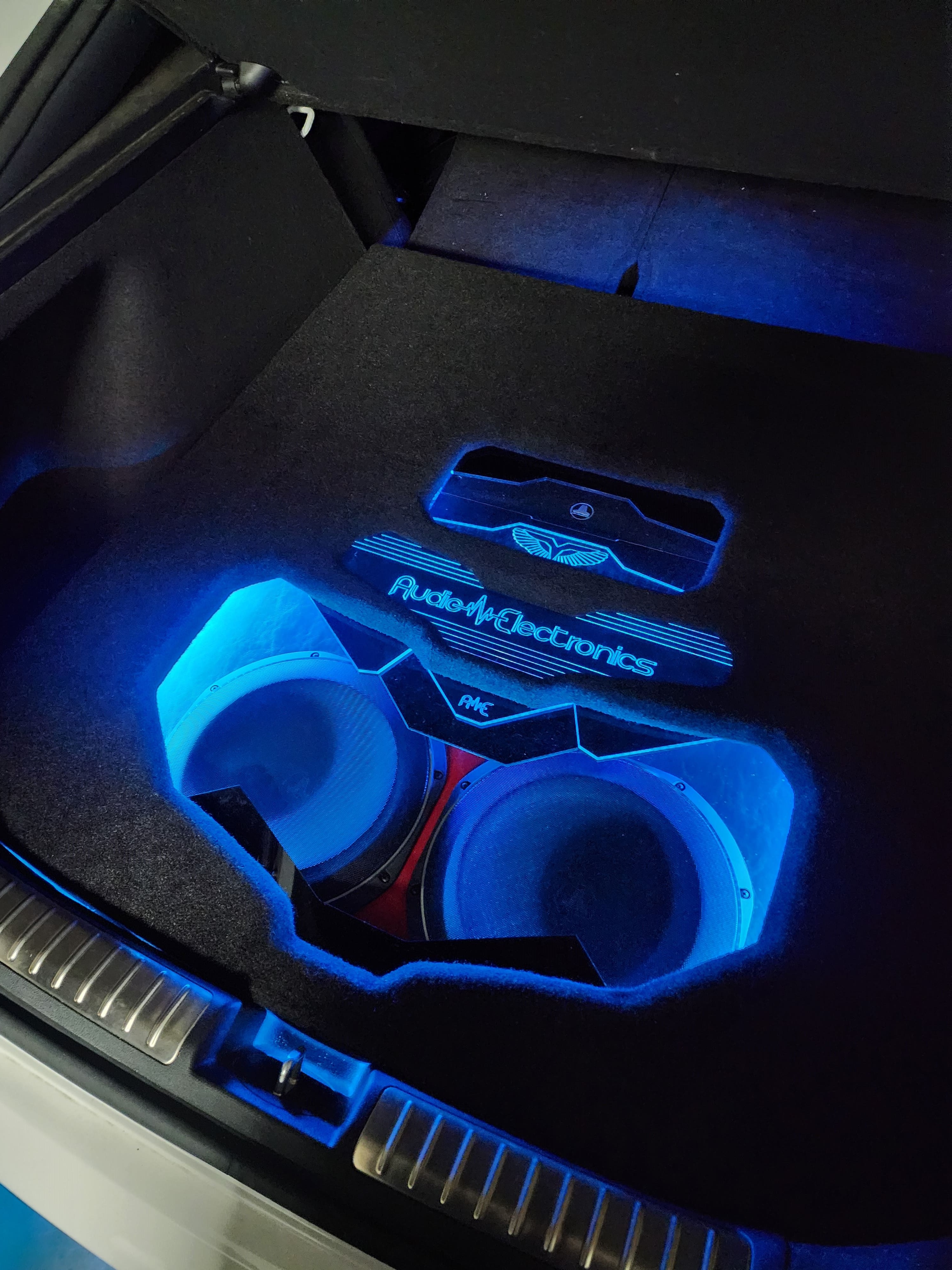 Two subwoofers in a carpeted trunk build, framed by blue accent lighting and a backlit Audio Electronics logo.