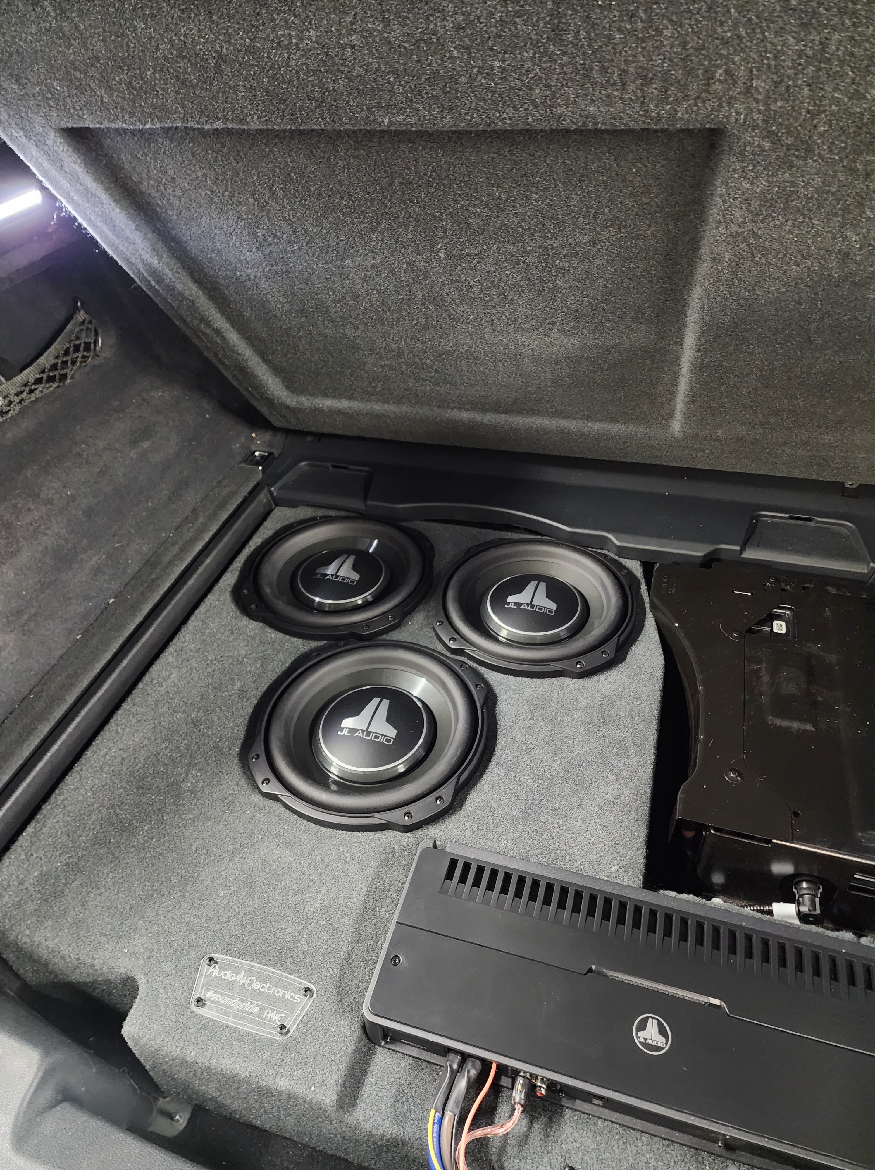 Three shallow mount subwoofers set into a carpeted trunk floor next to a mounted multichannel amplifier.