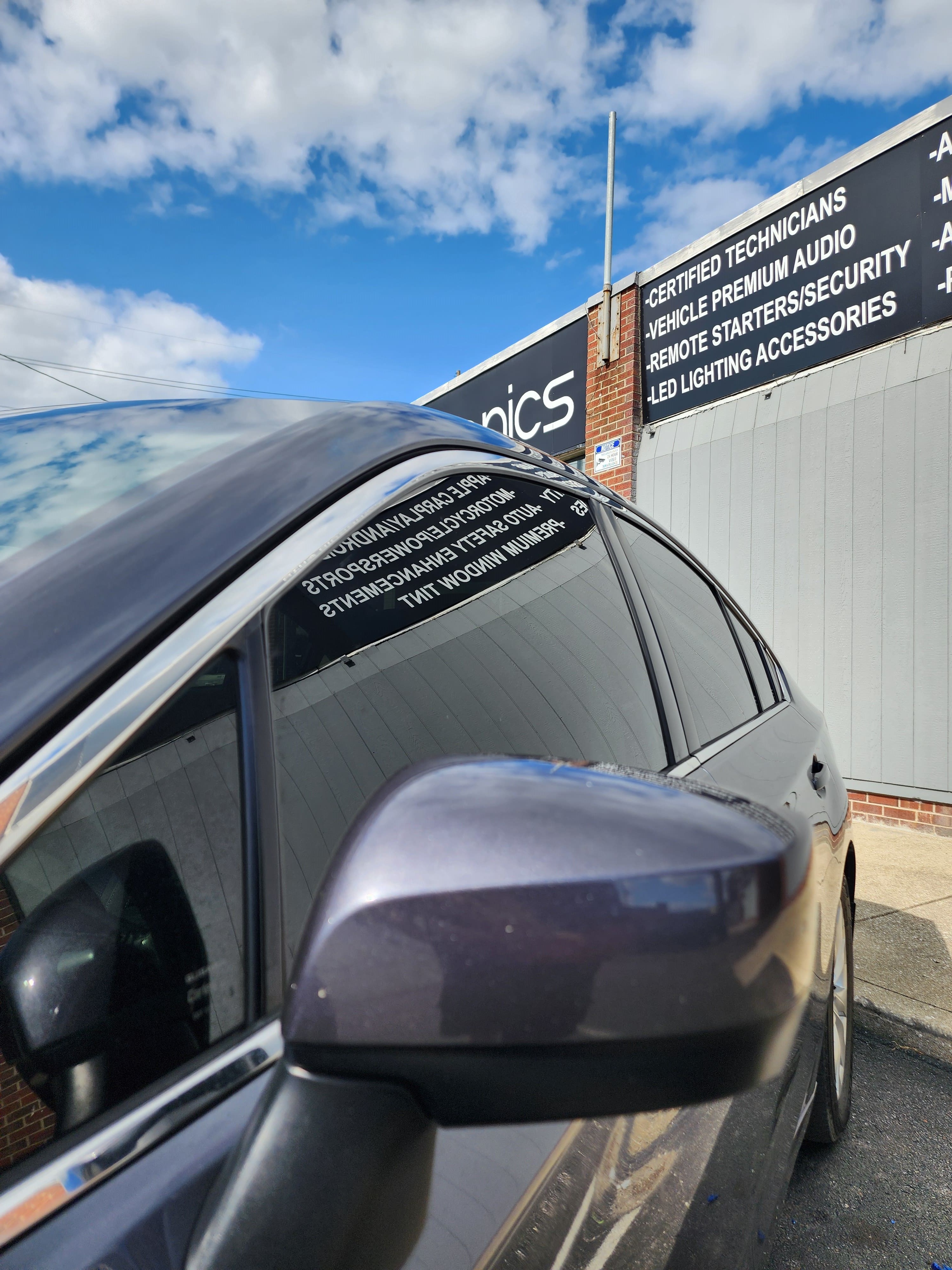 Dark tinted side windows on a gray sedan parked outside a shop with signage for audio, security and lighting services.