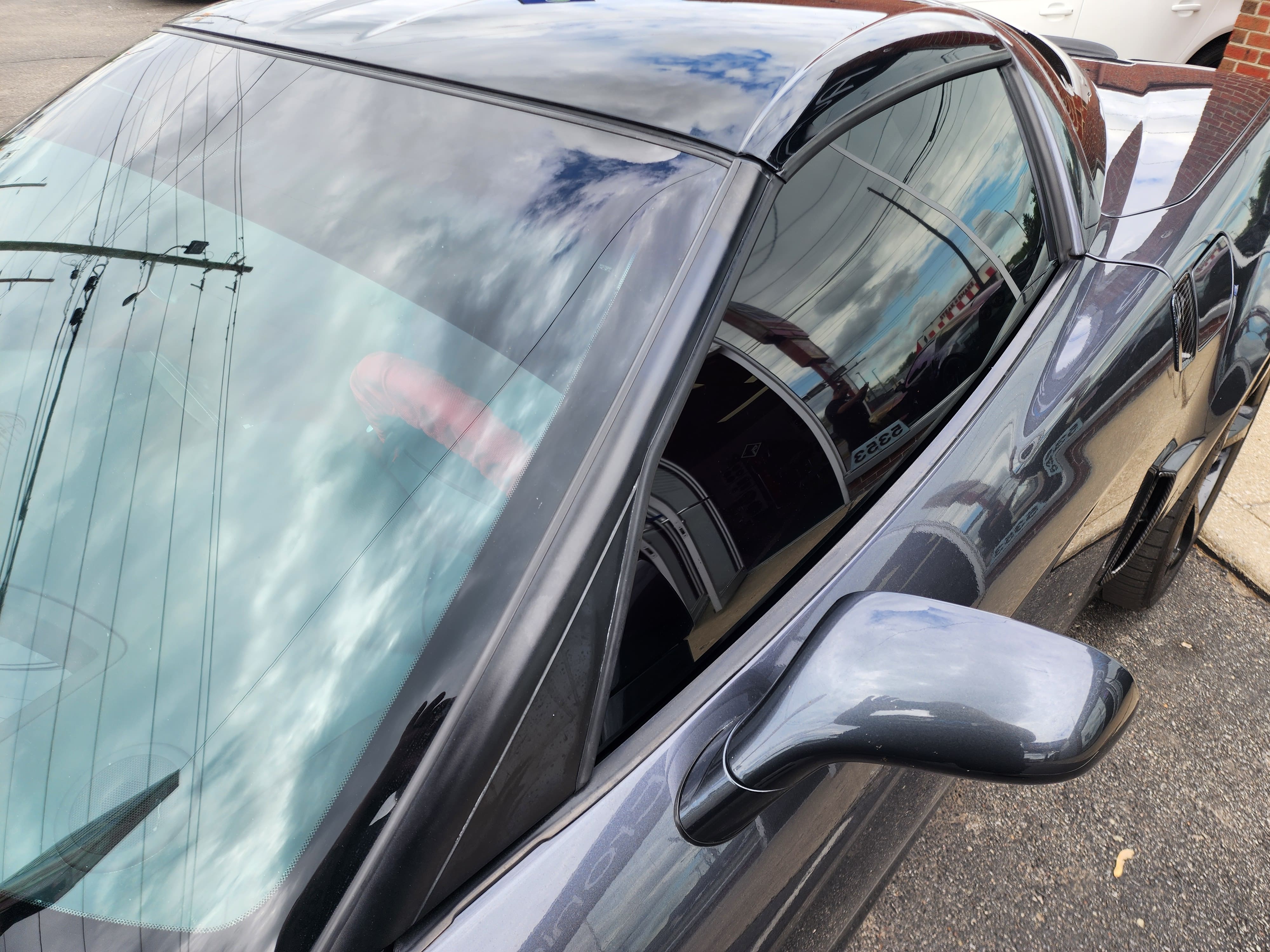 Close view of a dark sedan's tinted windshield and side glass reflecting a cloudy sky in a parking area.