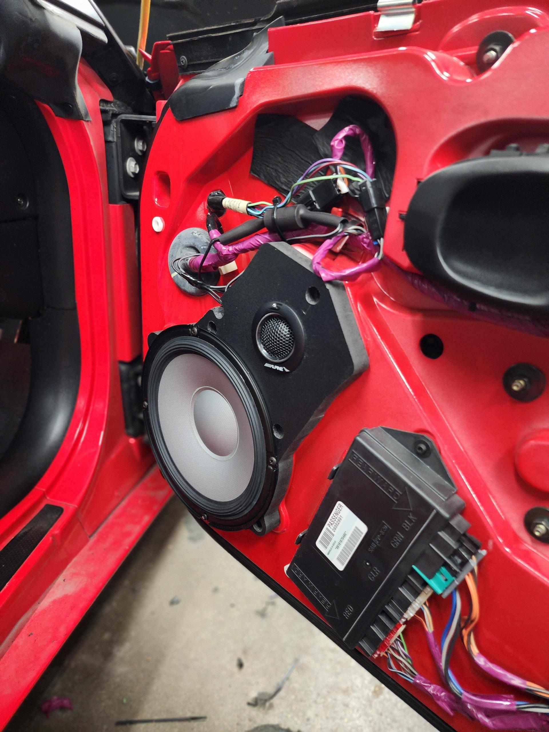 Red car door with the trim panel removed, showing a mounted woofer, tweeter, wiring harness and a control module.