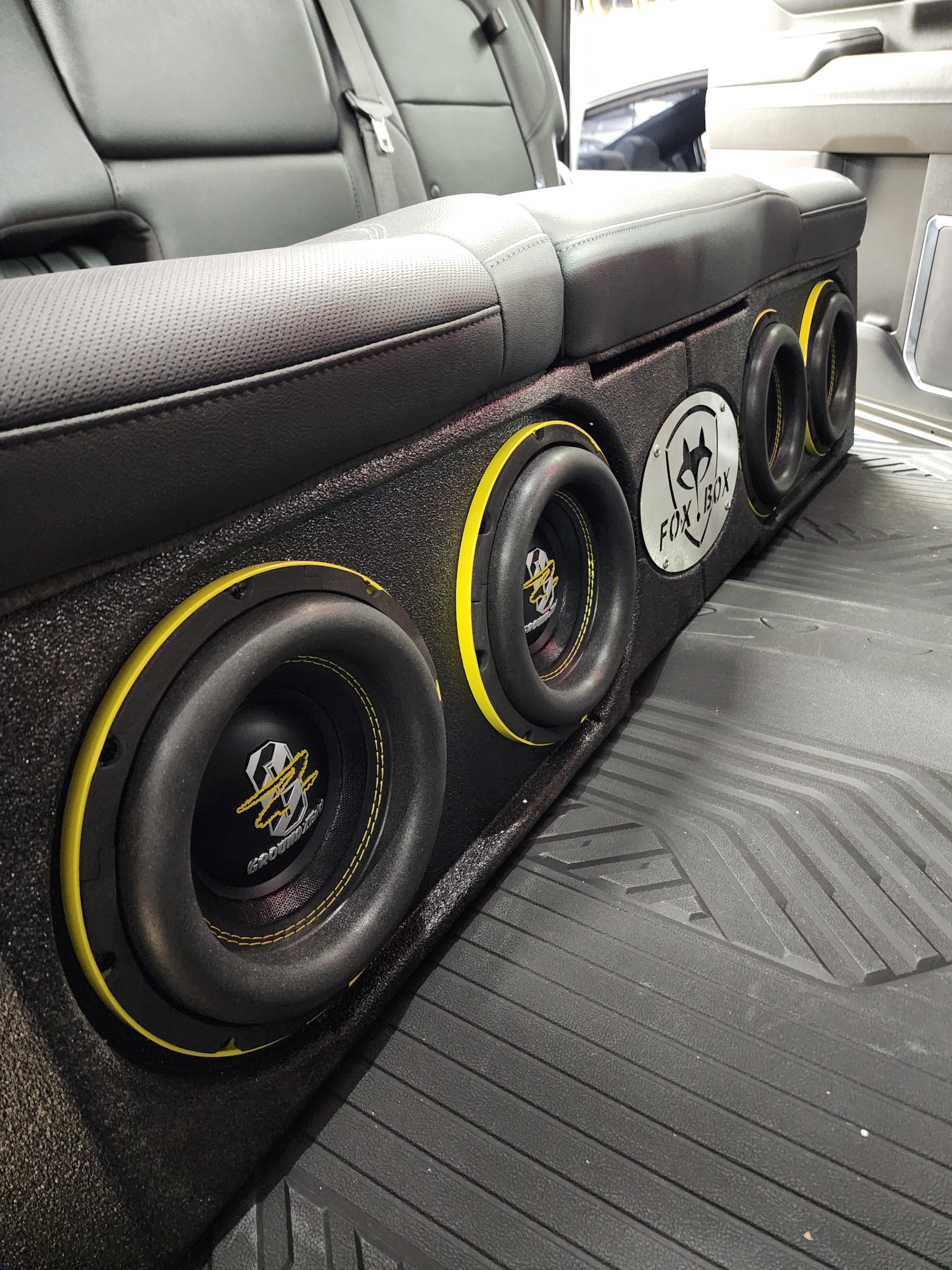 Four subwoofers mounted in a long carpeted enclosure fitted behind the rear seat of a pickup truck cab.