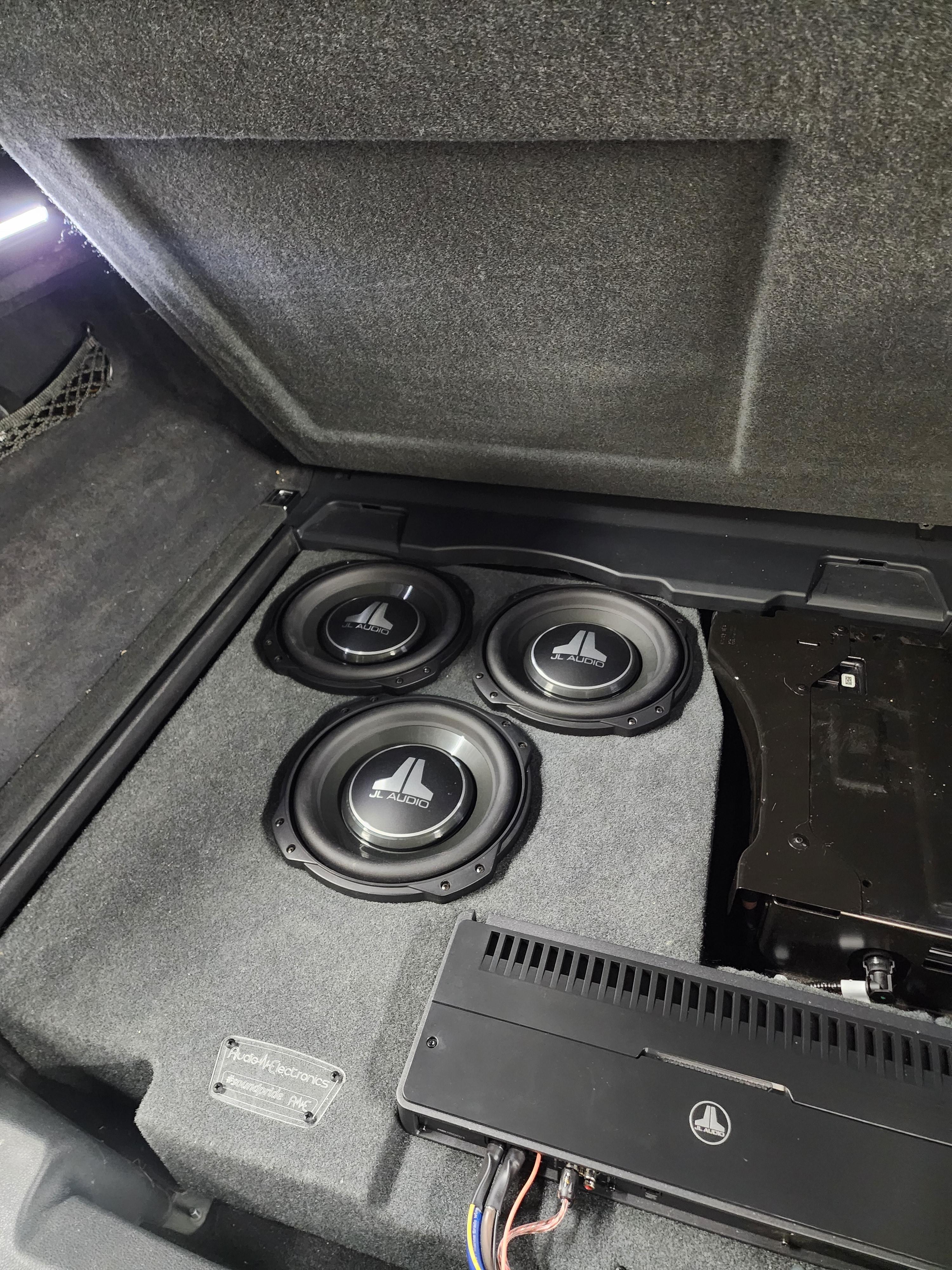 Three subwoofers flush mounted in a carpeted trunk panel beside a black amplifier with wiring running to it.