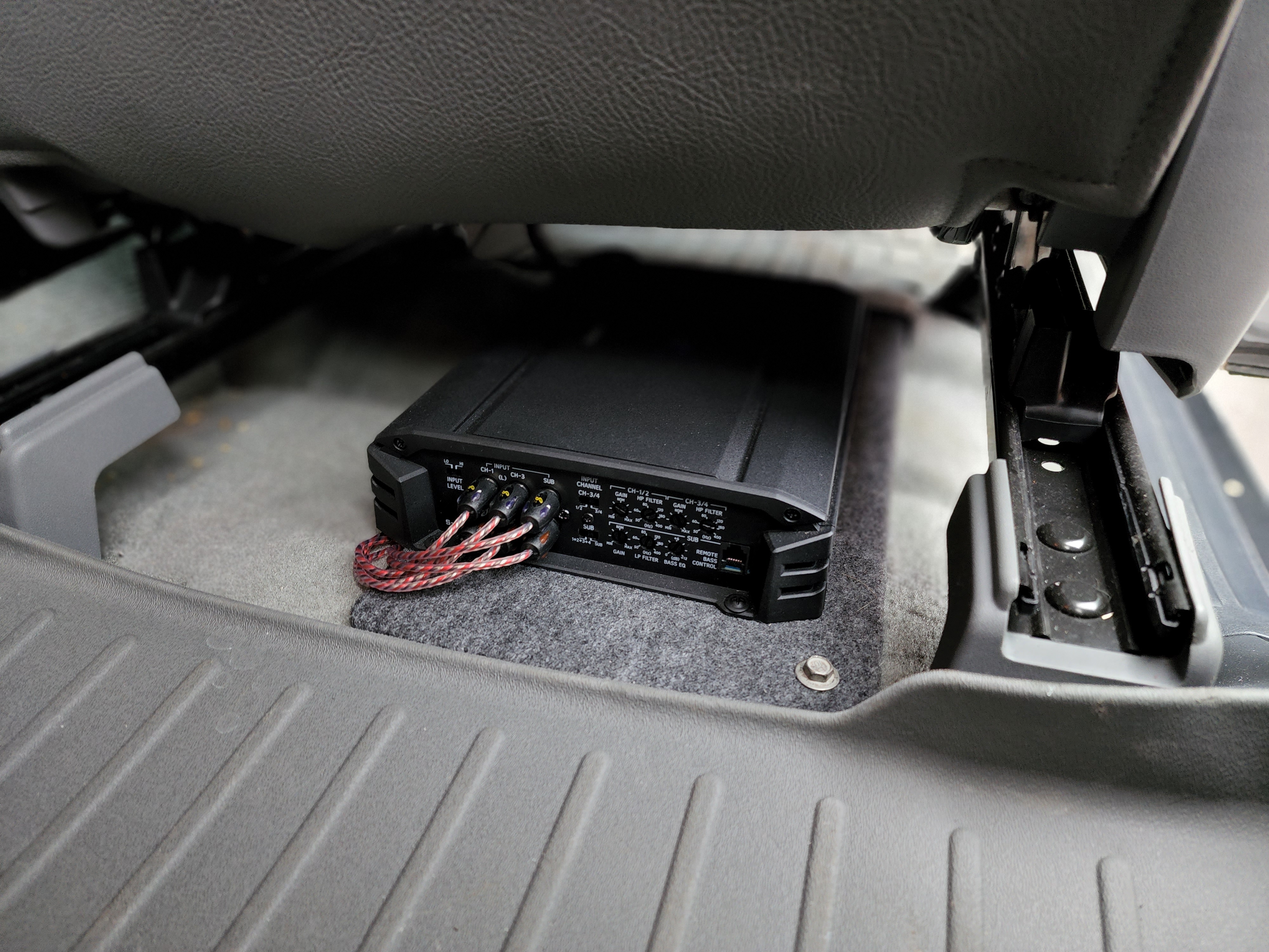 Compact car amplifier secured to the floor under a seat with red signal cables plugged into its inputs.