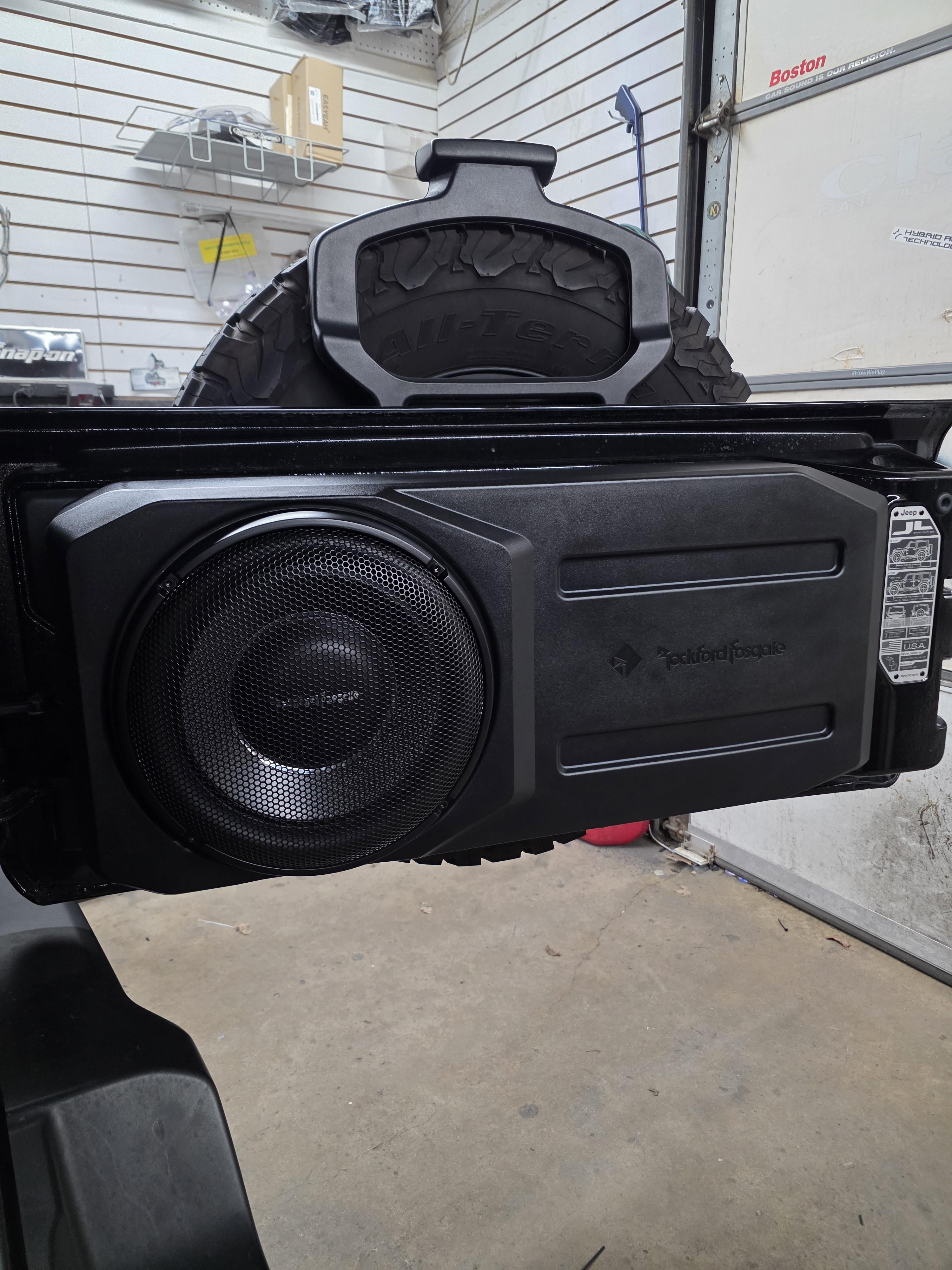 Slim loaded subwoofer enclosure with a mesh grille sitting in the open rear of a vehicle inside a shop.