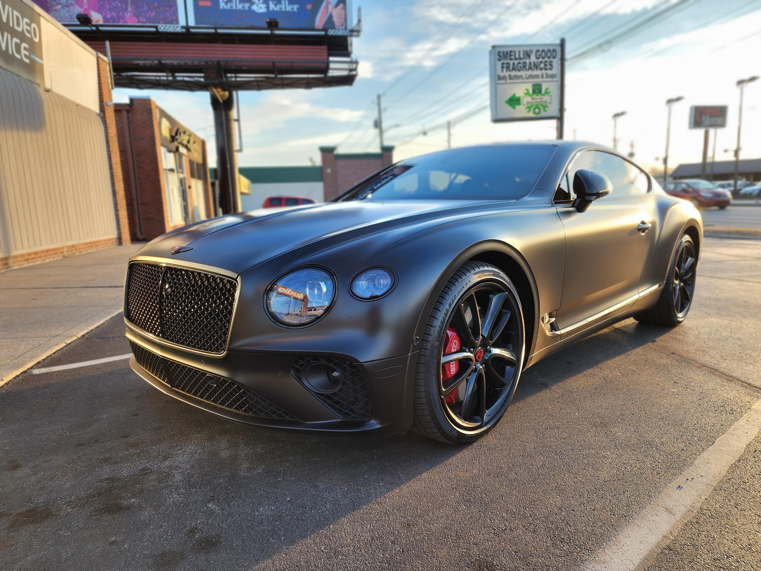 Matte black Bentley coupe with dark tinted windows and black wheels parked on pavement near a storefront.