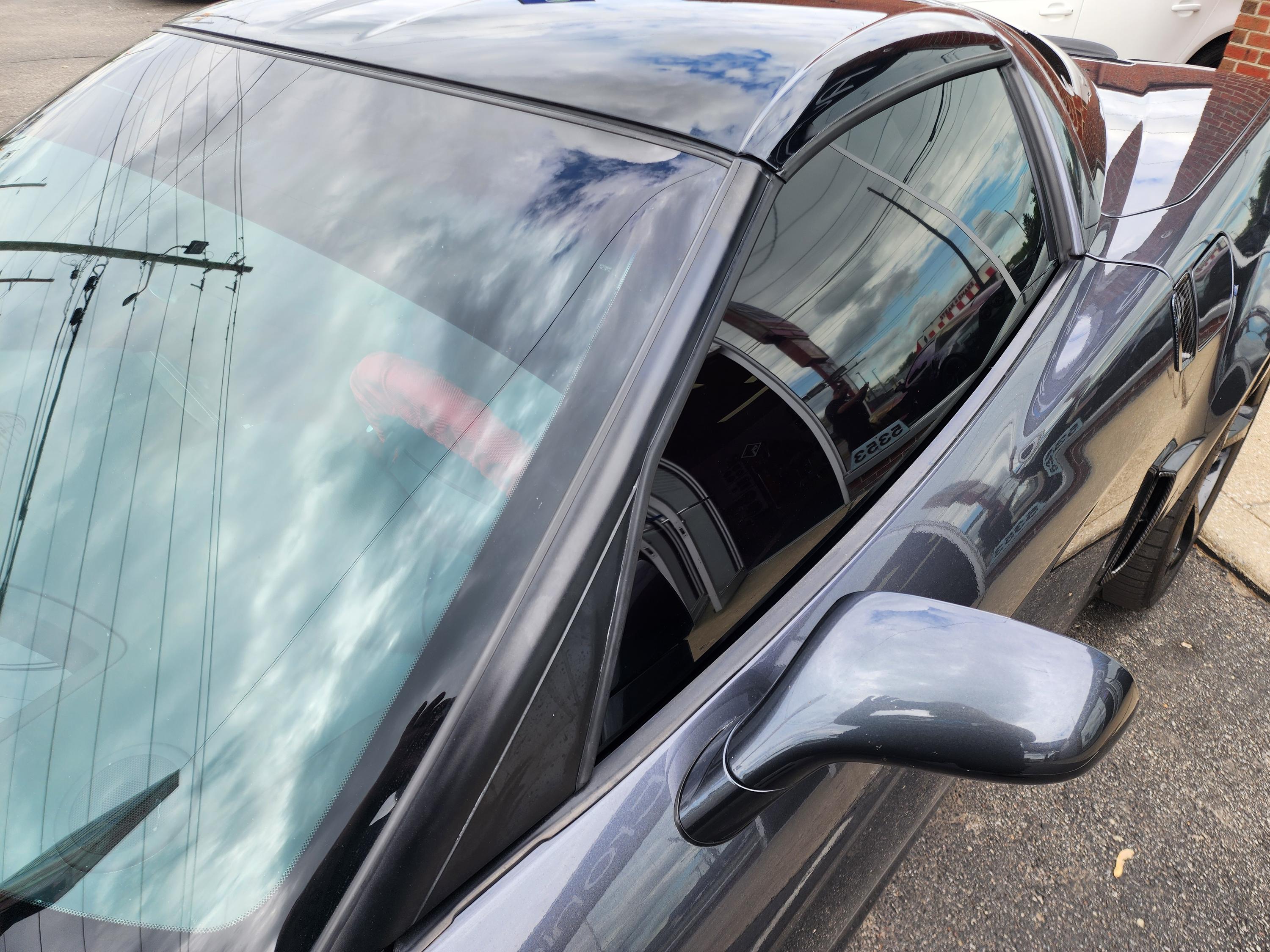 Close view of a dark car's windshield and tinted driver window reflecting clouds and overhead power lines.