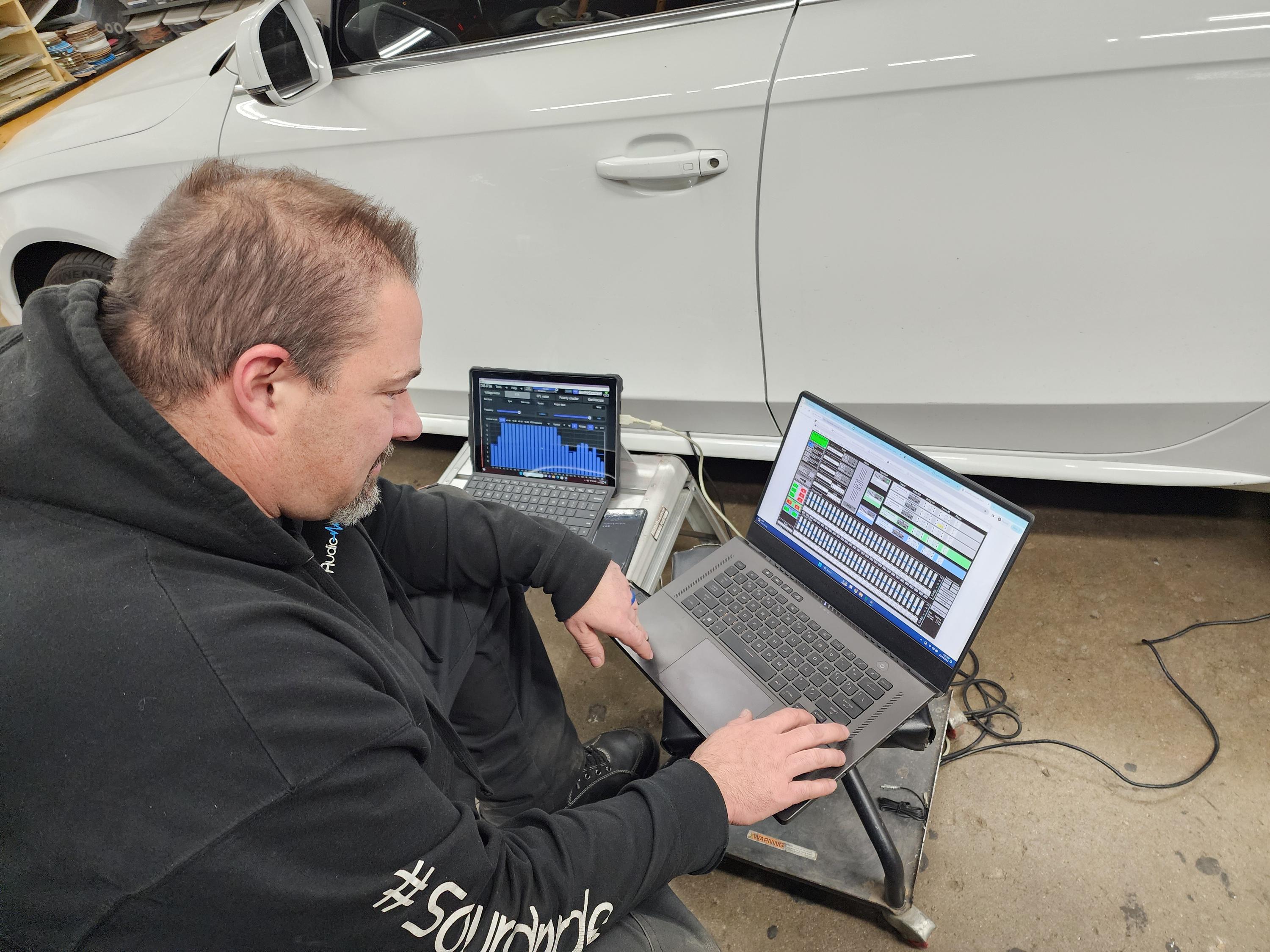 Installer tuning a car audio system on a laptop and tablet next to a white sedan.