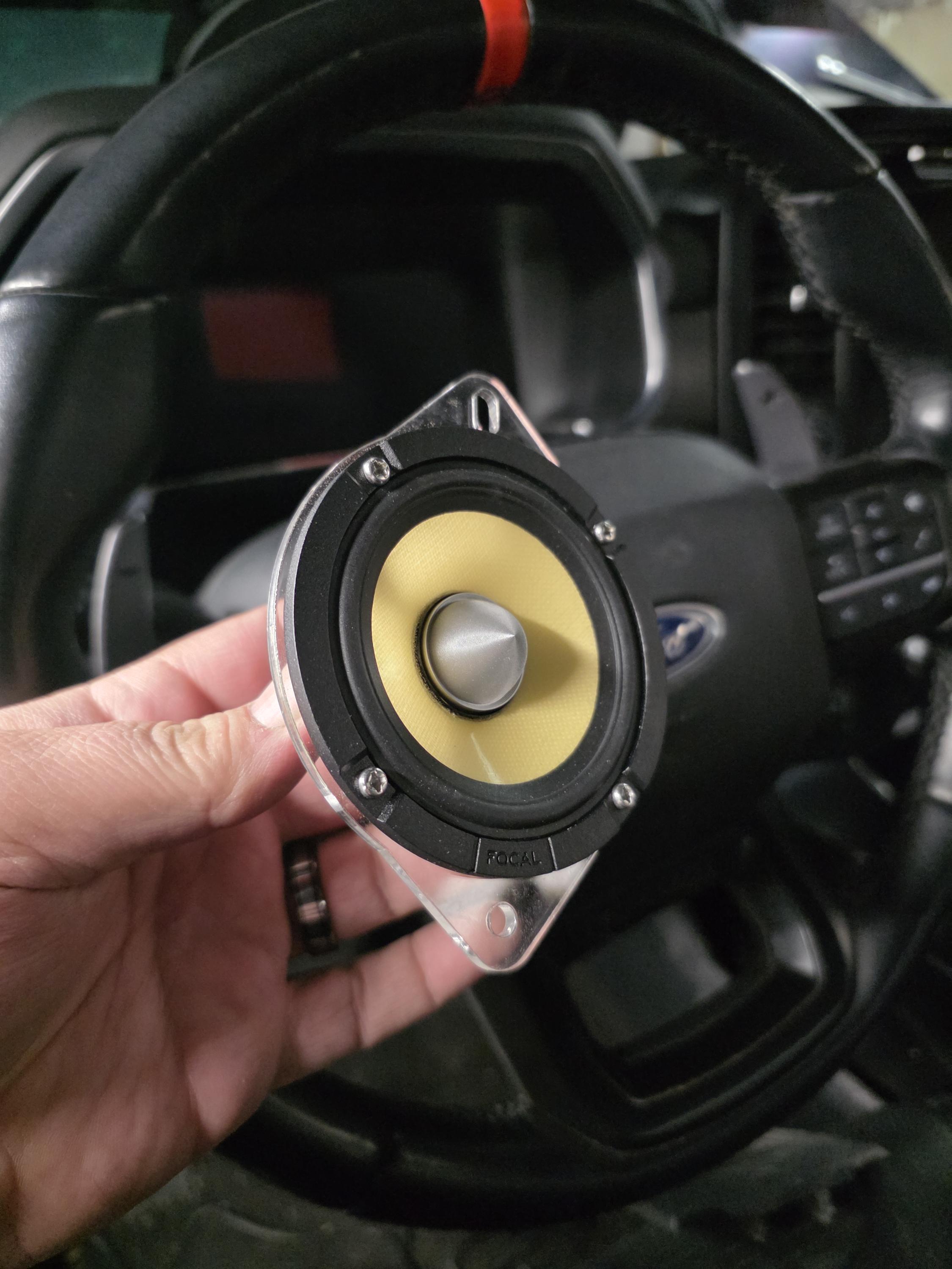 Hand holding a yellow cone speaker on a metal mounting bracket in front of a steering wheel