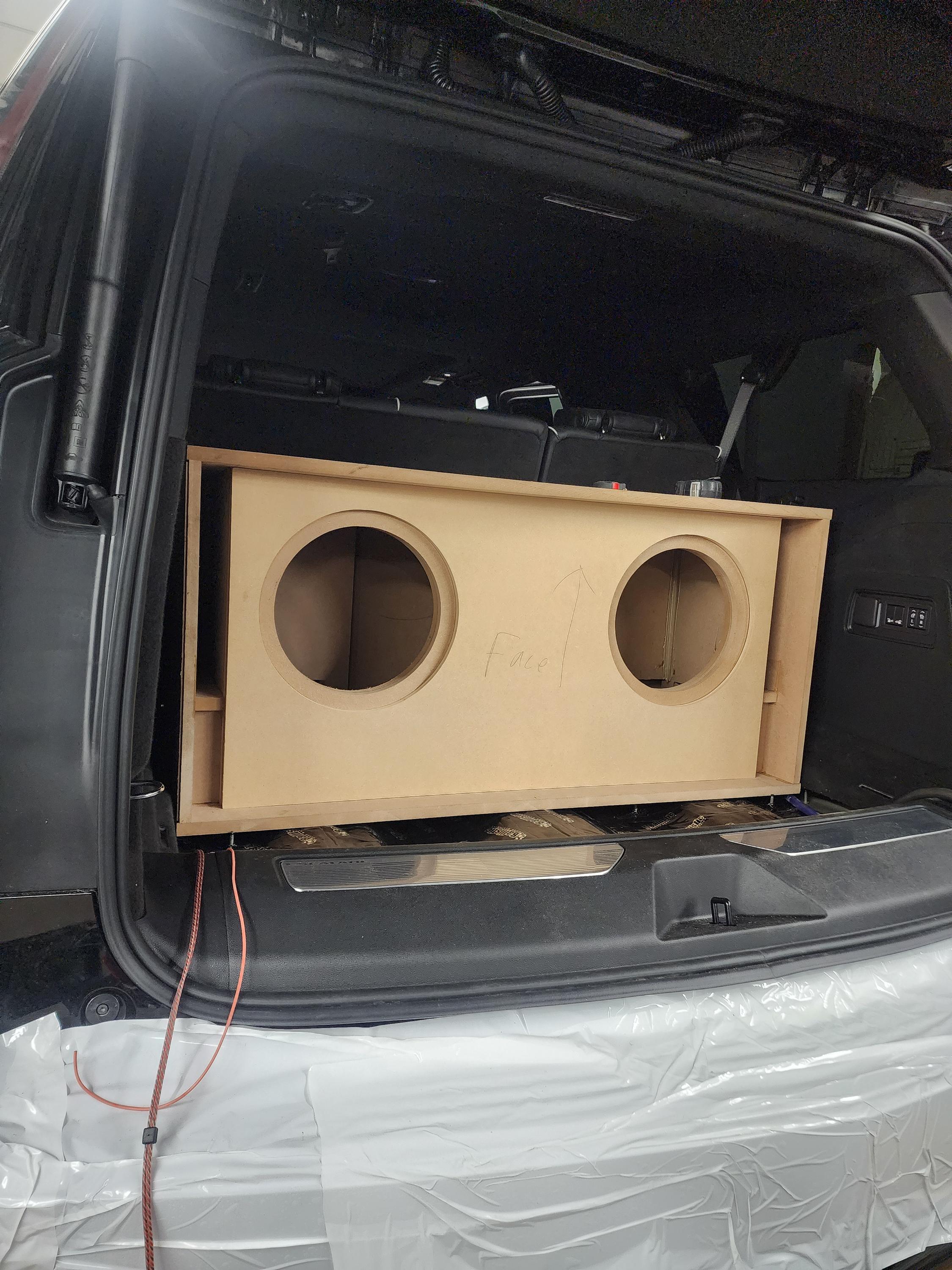 Unfinished wood subwoofer enclosure with two round cutouts test fitted in the cargo area of an SUV
