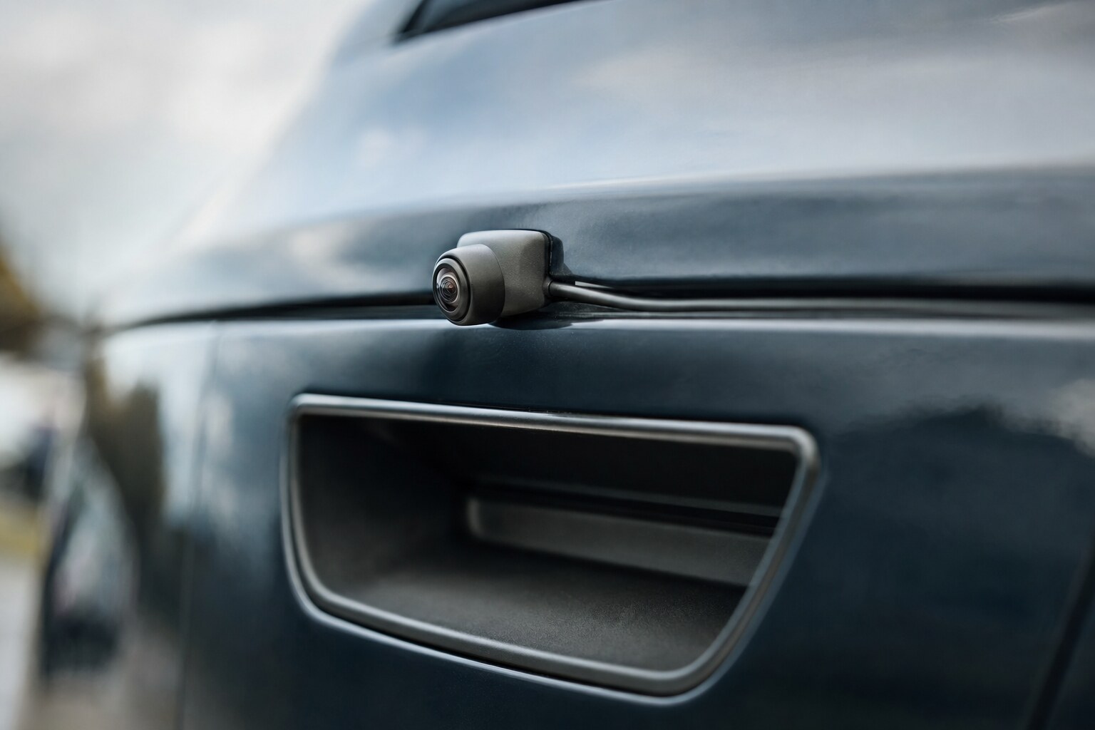 Small black backup camera and its cable mounted on the rear trim panel of a car just above the tailgate handle.