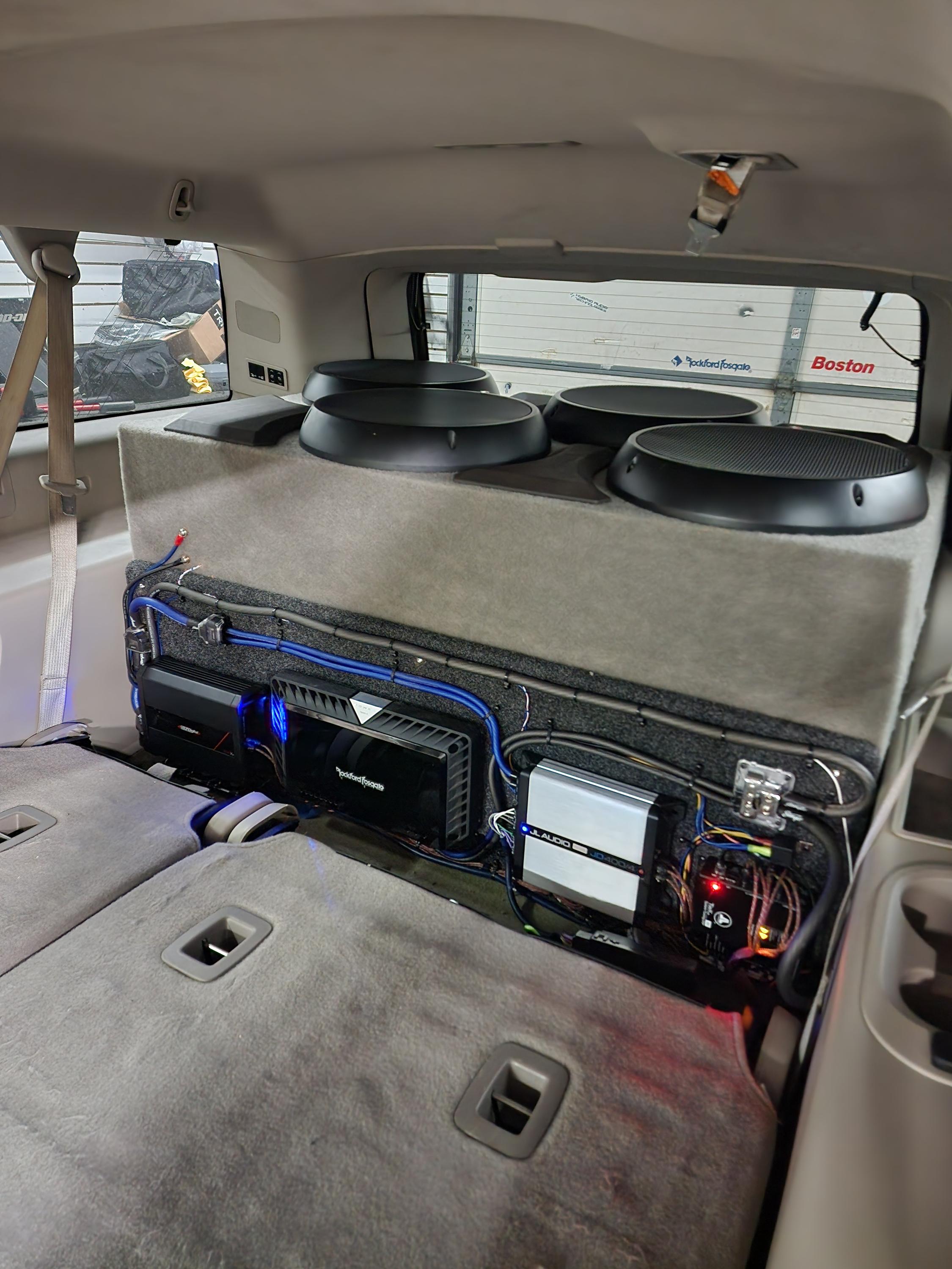 Four upward facing subwoofers on a carpeted deck above mounted amplifiers and wiring in an SUV cargo area