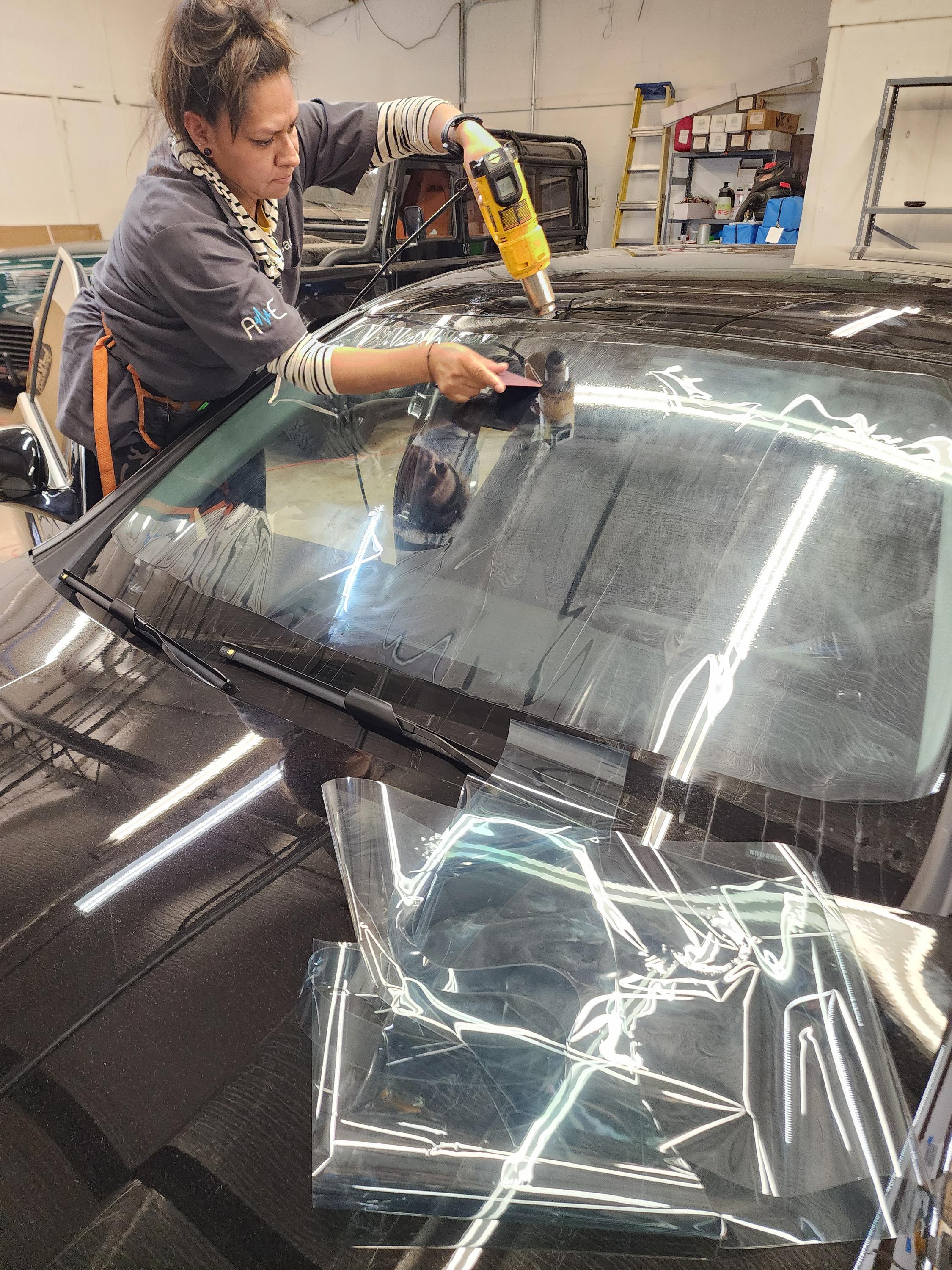 Installer uses a heat gun and a squeegee to shrink and shape window film across a car windshield indoors.