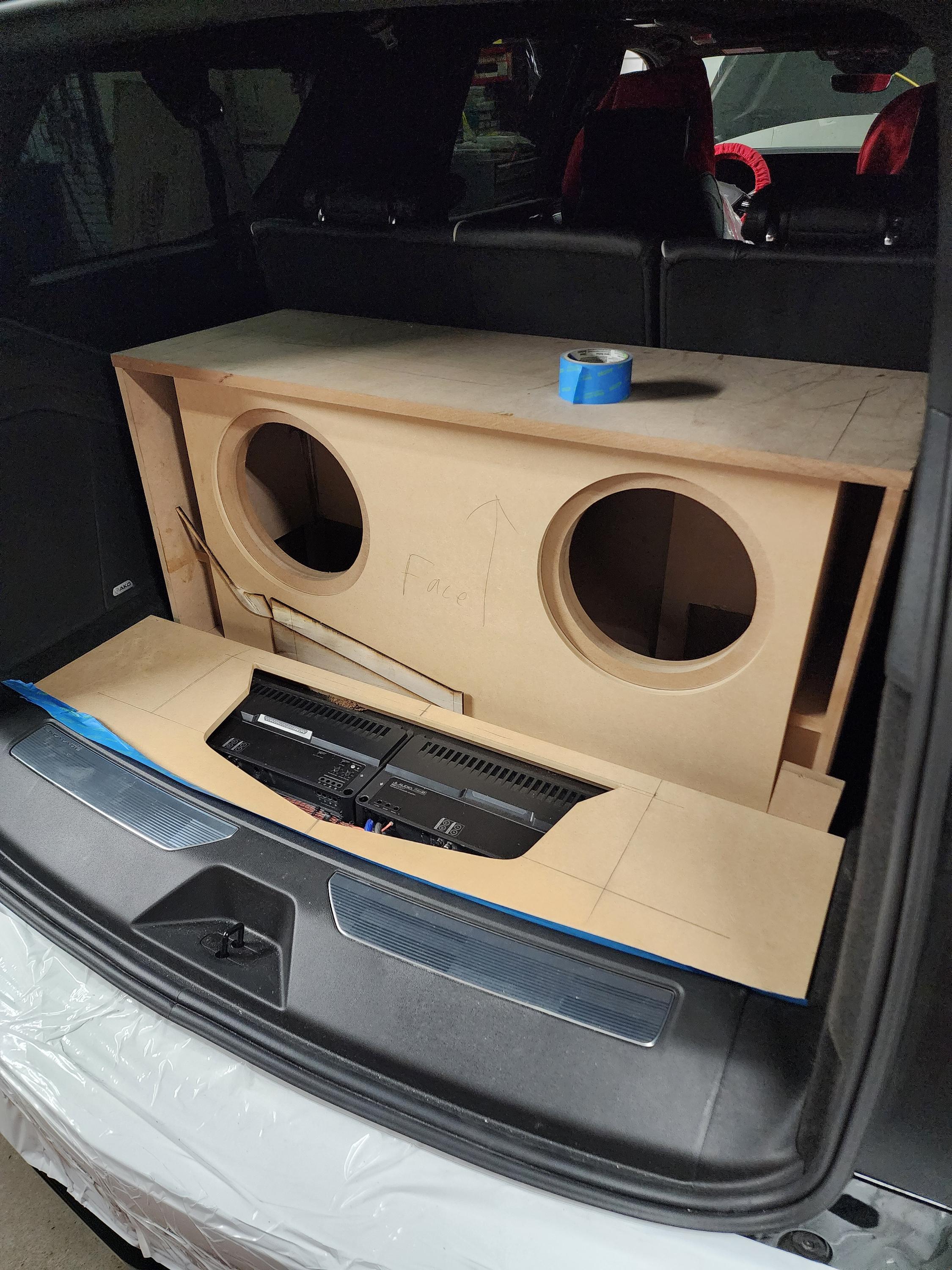 Unfinished wood subwoofer enclosure with two cutouts and an amplifier board test fitted in a vehicle cargo area.